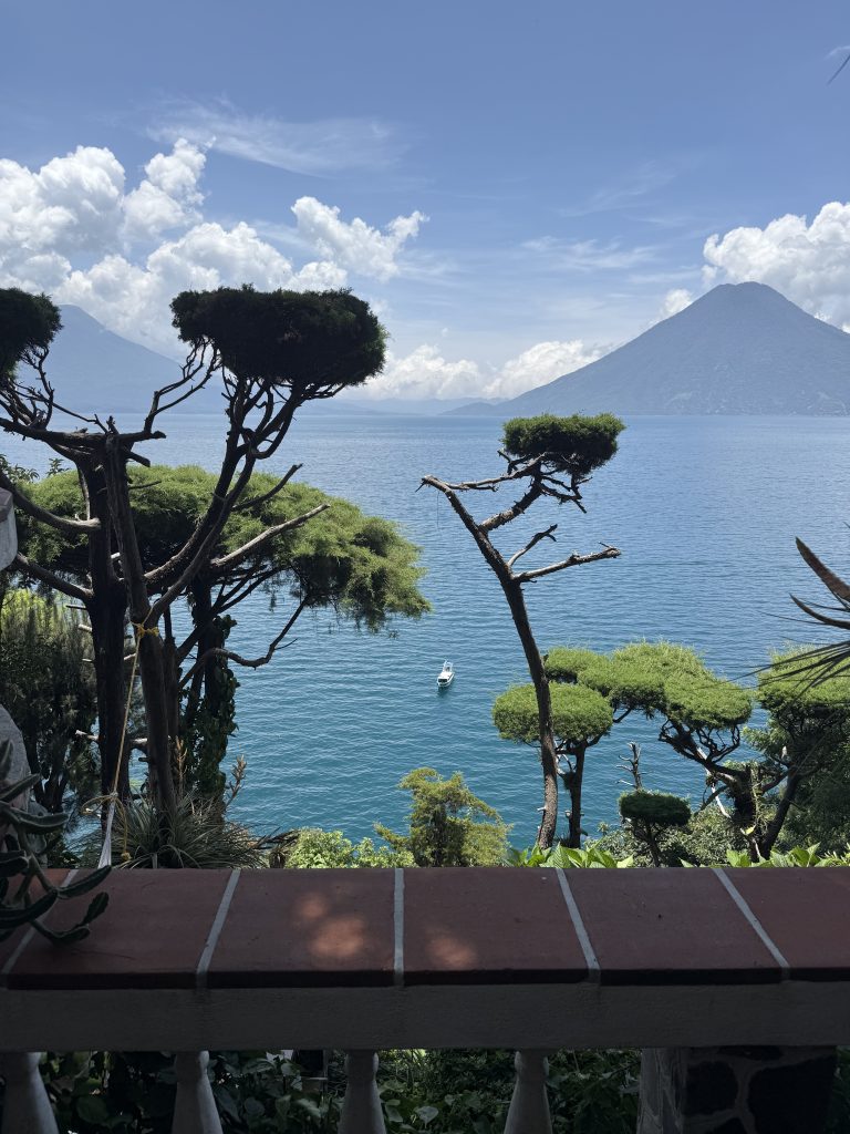 View from a terrace above vivid blue Lake Atitlán with sculpted trees and twin volcanoes on a humid rainy-season day.