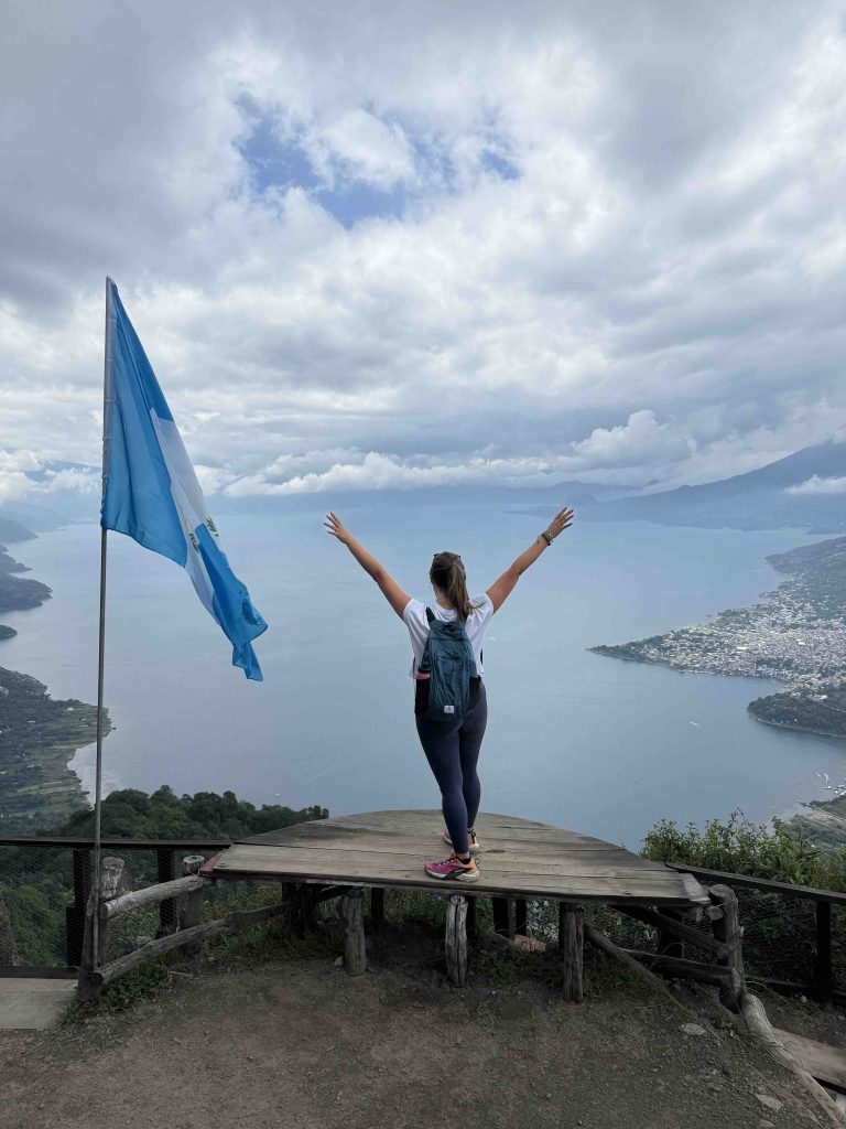 Woman standing with arms raised at the Indian Nose summit overlooking Lake Atitlán and villages below