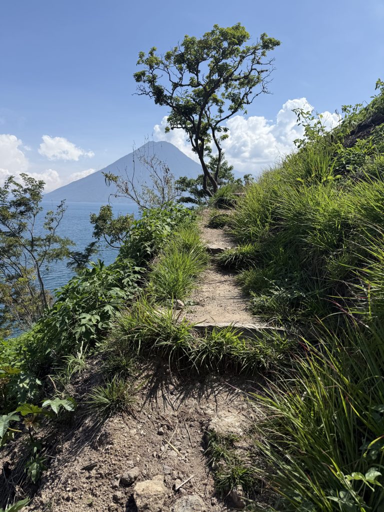 Narrow lakeside hiking trail on grassy ridge with volcano in background
