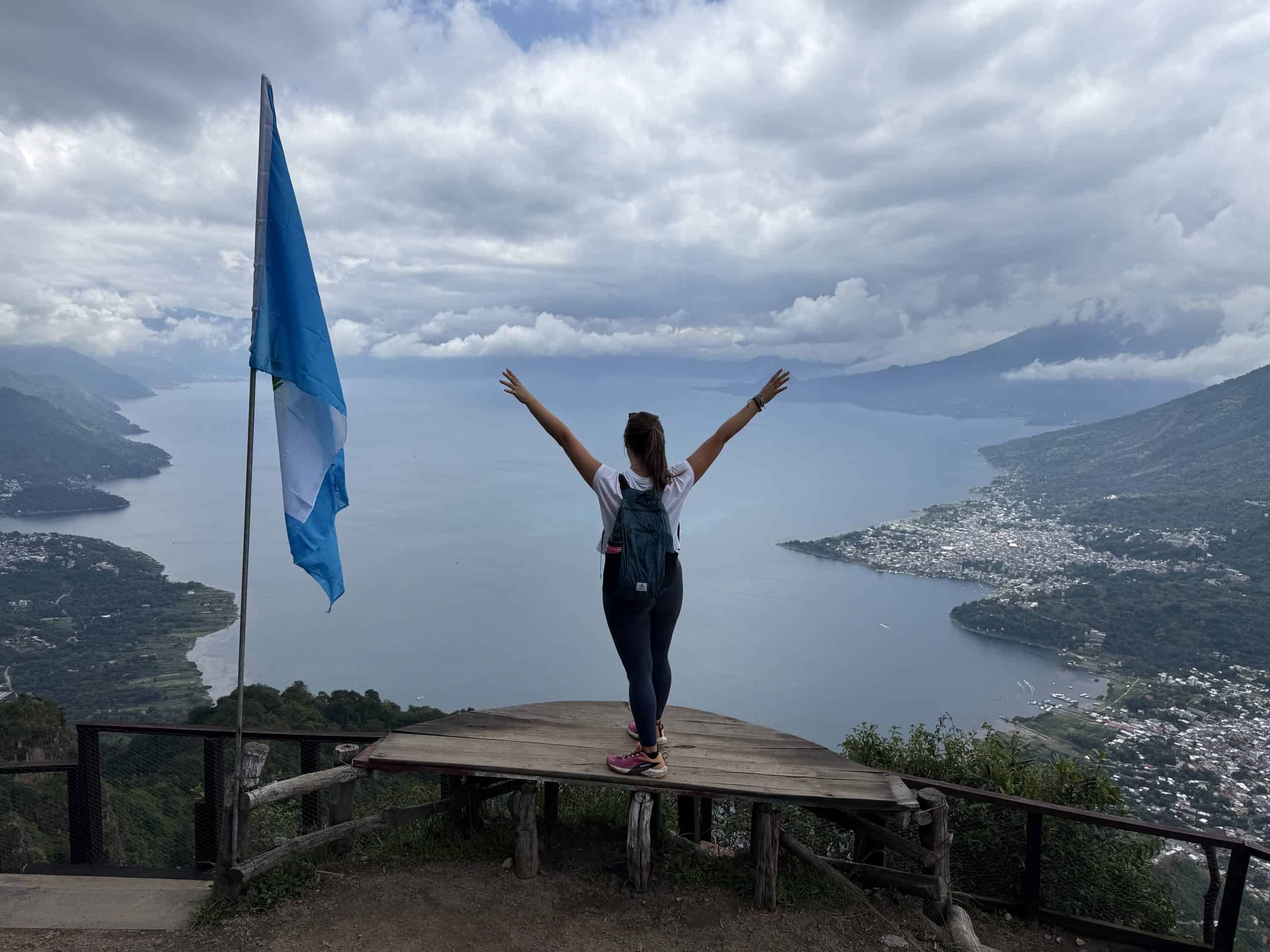 Hiker with arms raised at Lake Atitlán viewpoint next to Guatemalan flag