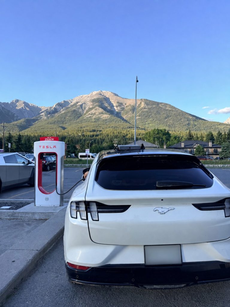 Ford Mustang Mach-E charging at a Tesla Supercharger in Canmore, Alberta, with the Canadian Rockies in the background.