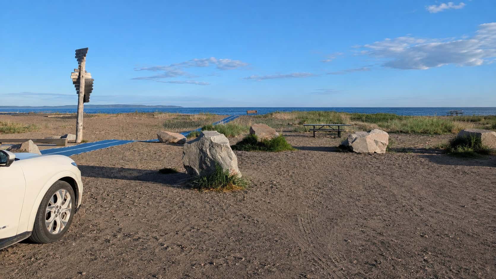 Scenic EV road trip stop along Lake Superior with a parked vehicle, rocky shoreline, picnic table, and blue sky.