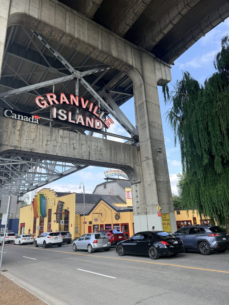 Granville Island entrance sign beneath the bridge with colourful buildings in Vancouver.