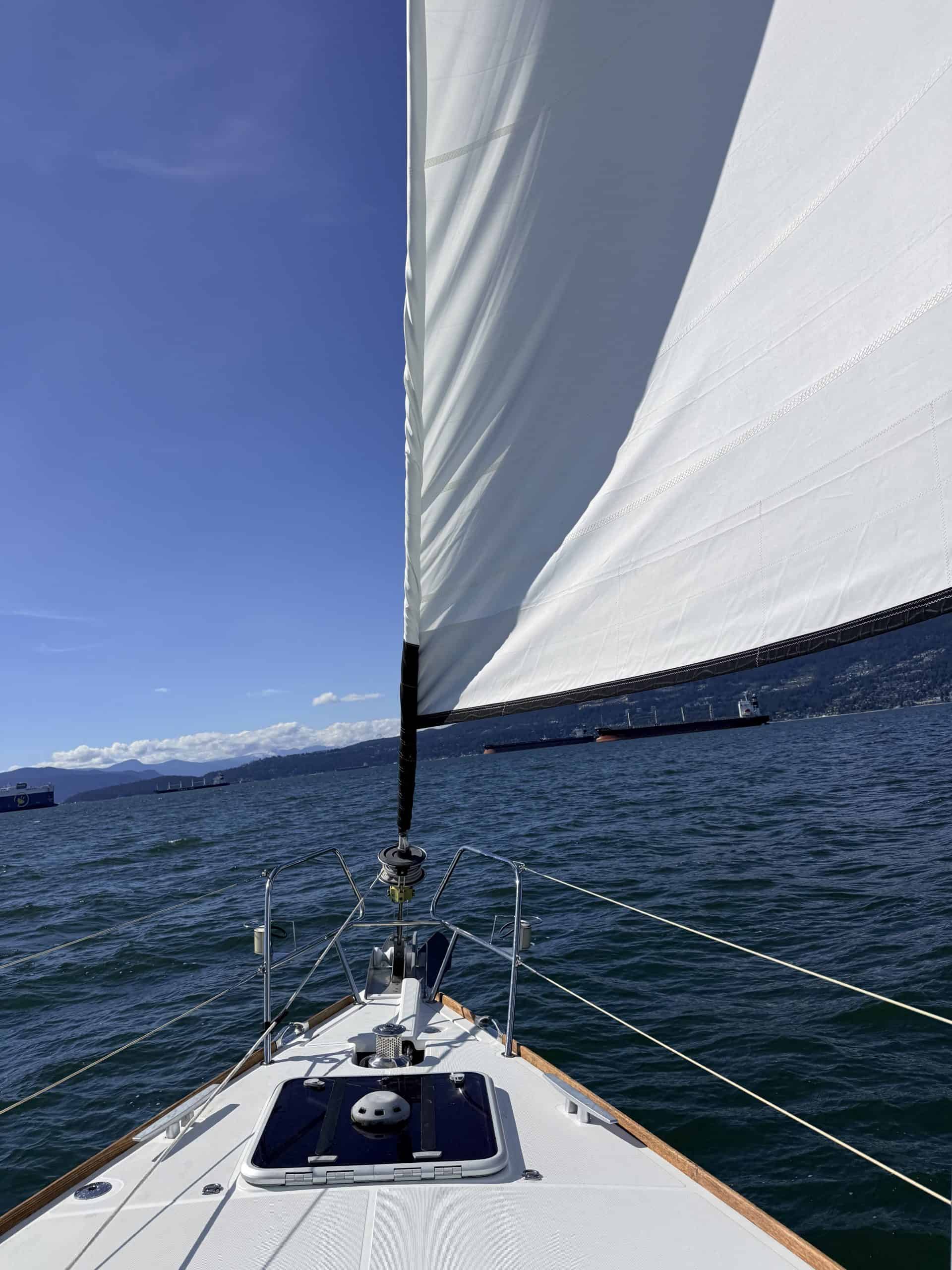 View from a sailboat in English Bay, Vancouver, with white sails, cargo ships and mountains in the distance.