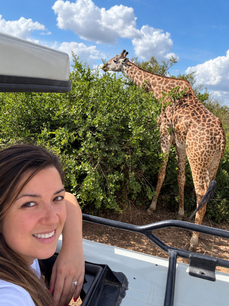 Annie smiling during a safari beside a giraffe under a bright blue sky.