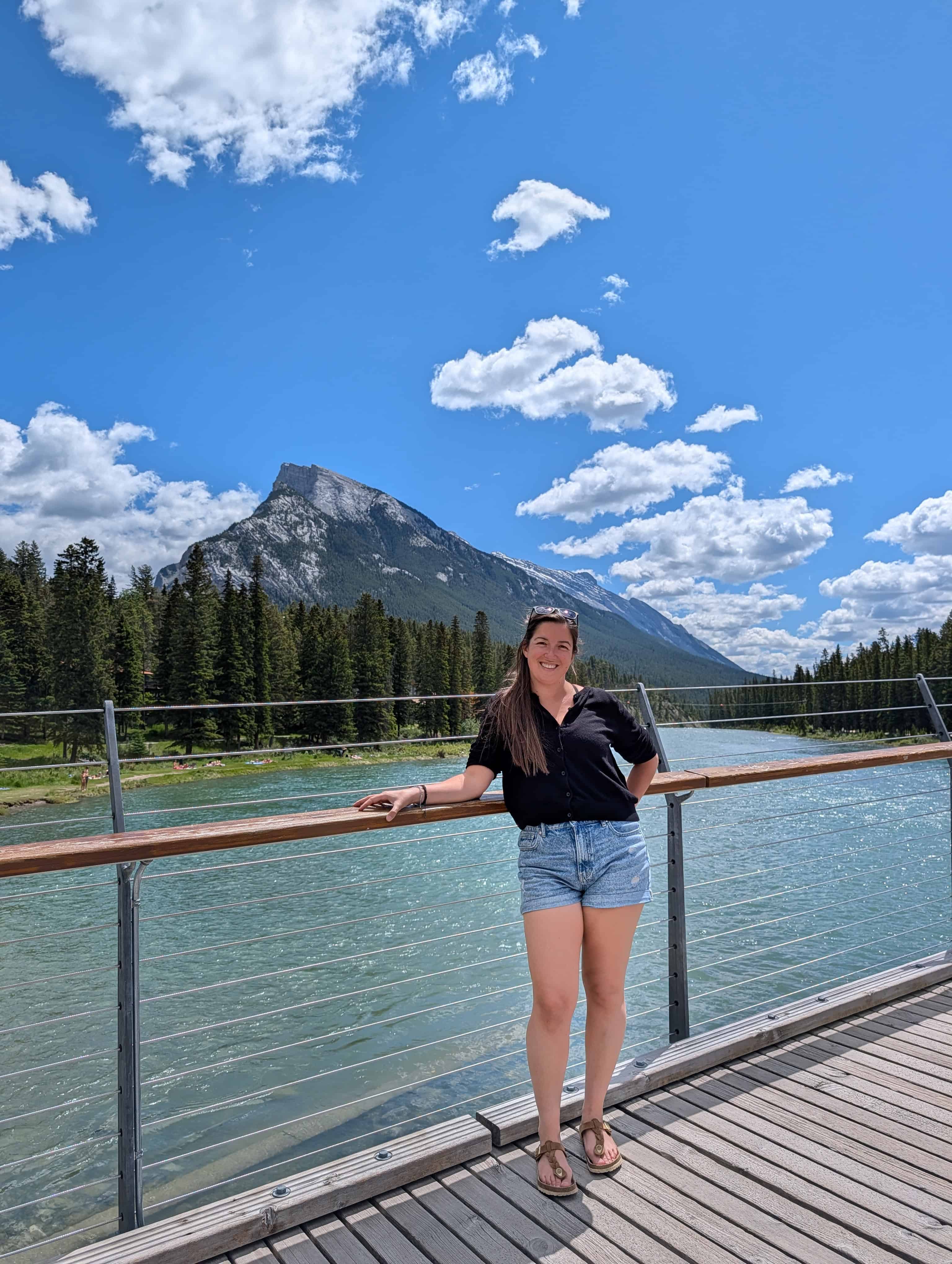 Annie standing on a pedestrian bridge over the turquoise Bow River in Banff, with forested mountains beneath a blue summer sky.