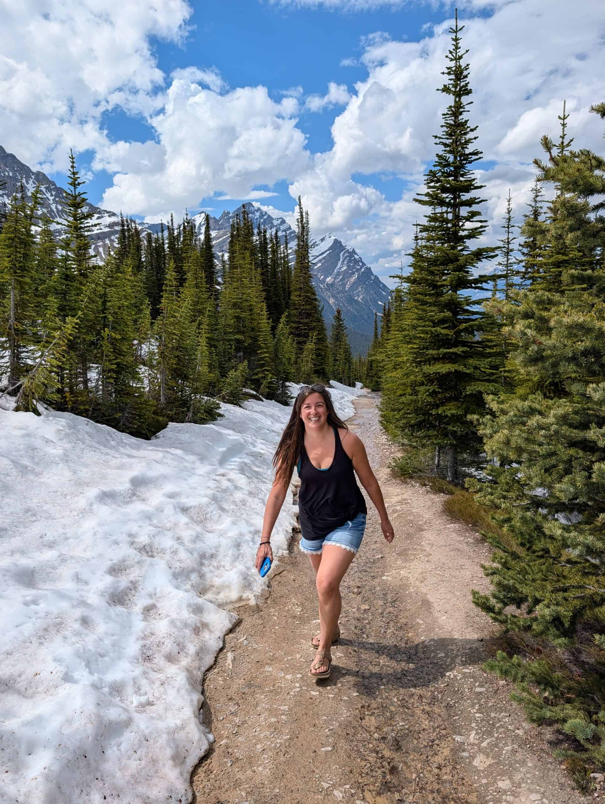 Annie walking along a gravel section of the Bow Summit trail beside lingering snowbanks and evergreen trees.