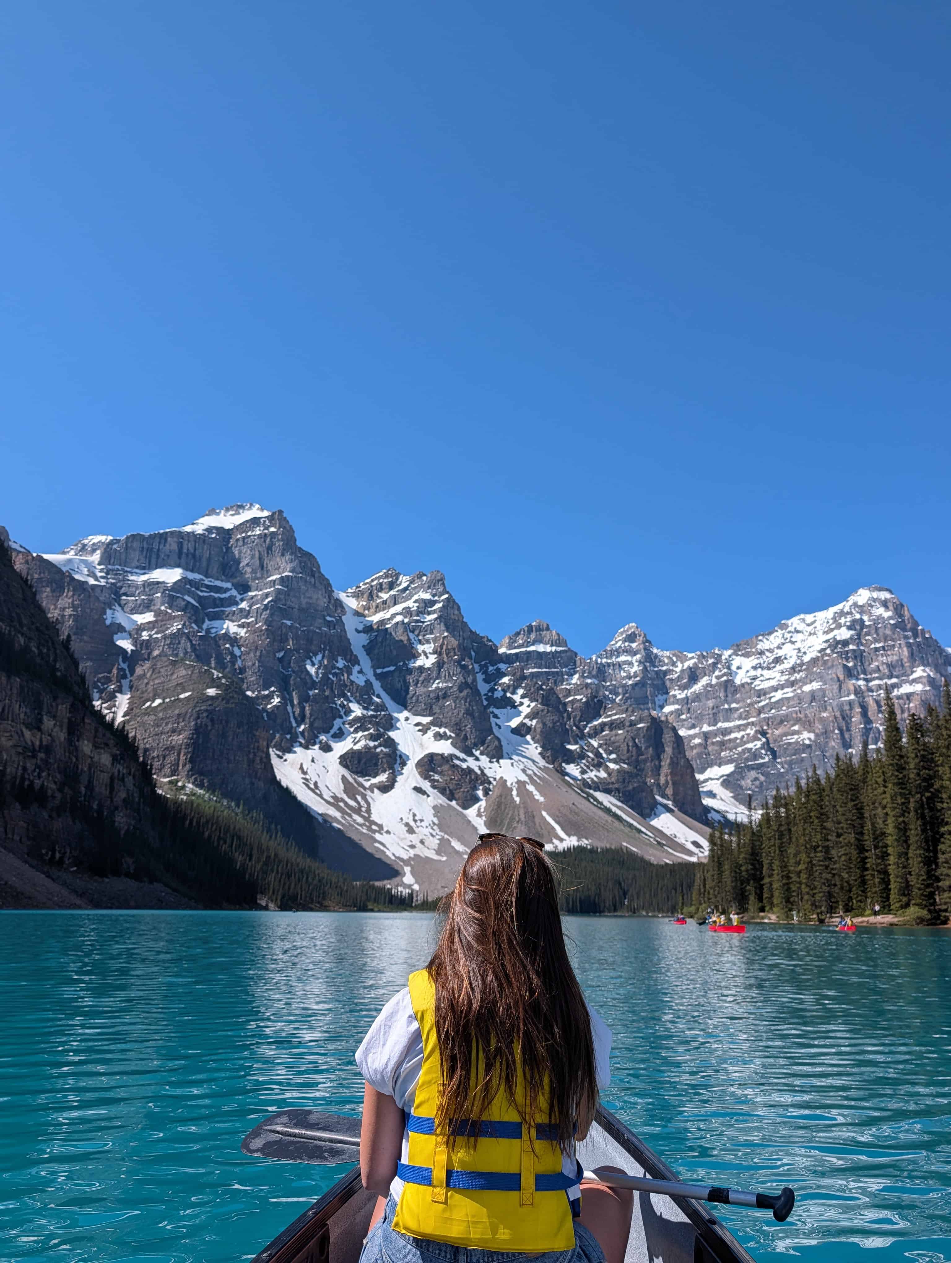 Annie wearing a yellow life jacket in a canoe on turquoise Moraine Lake, facing snow-covered peaks in Banff National Park.