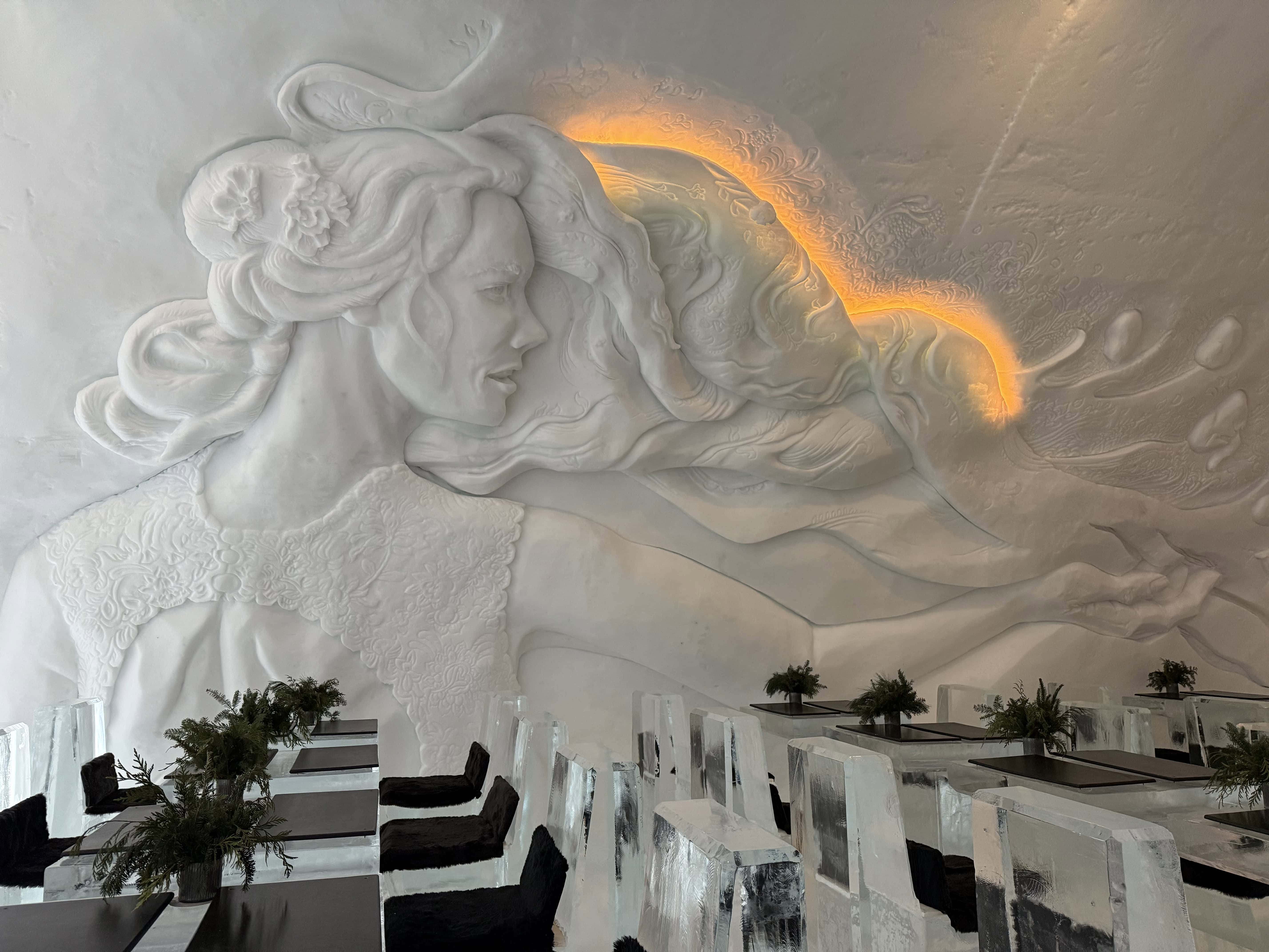 Ice dining room at Hôtel de Glace near Quebec City, with sculpted ice chairs, tables, and an illuminated wall carving.