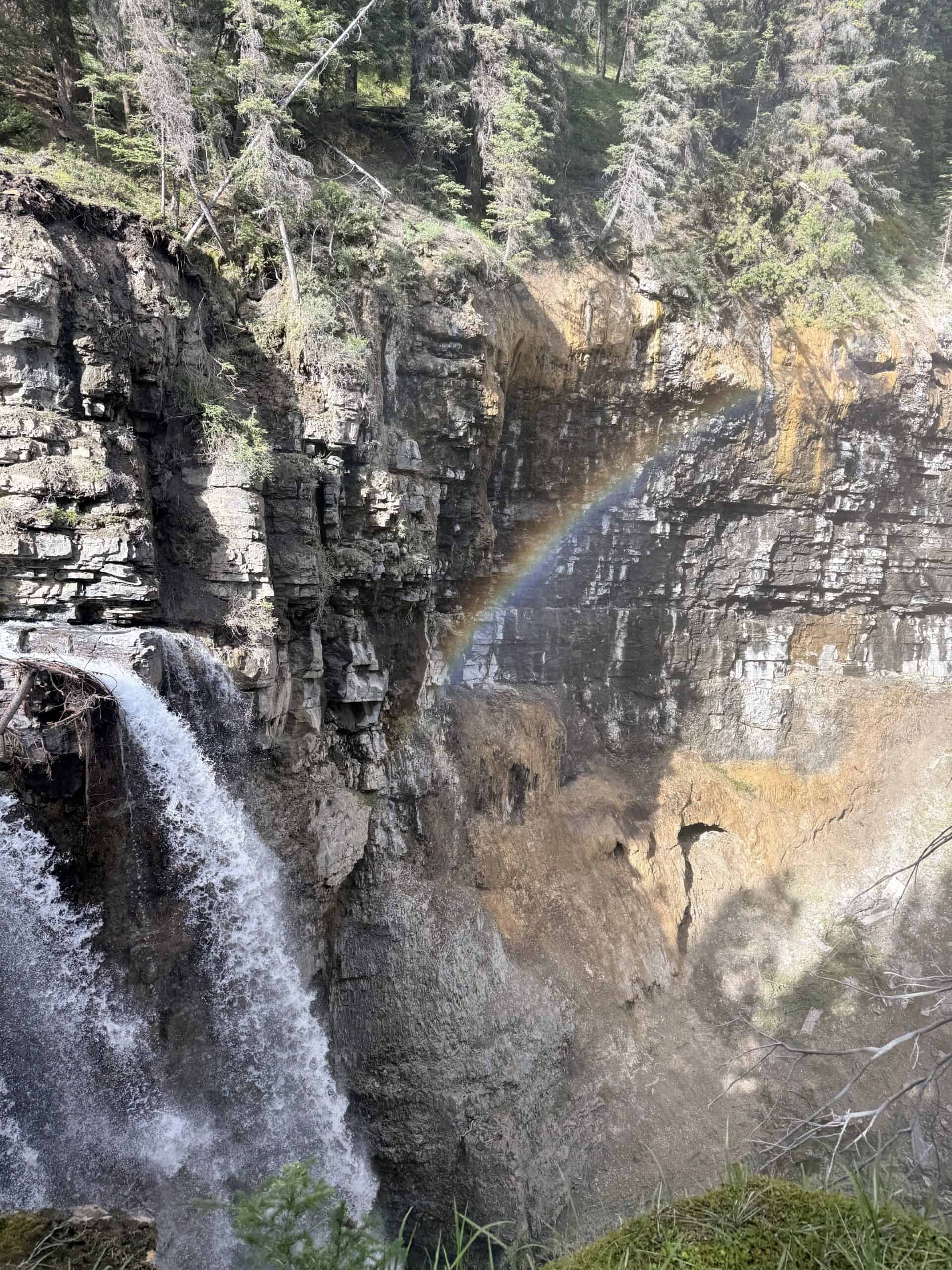 Rainbow forming in the mist beside a waterfall flowing through the rocky walls of Johnston Canyon in Banff National Park.