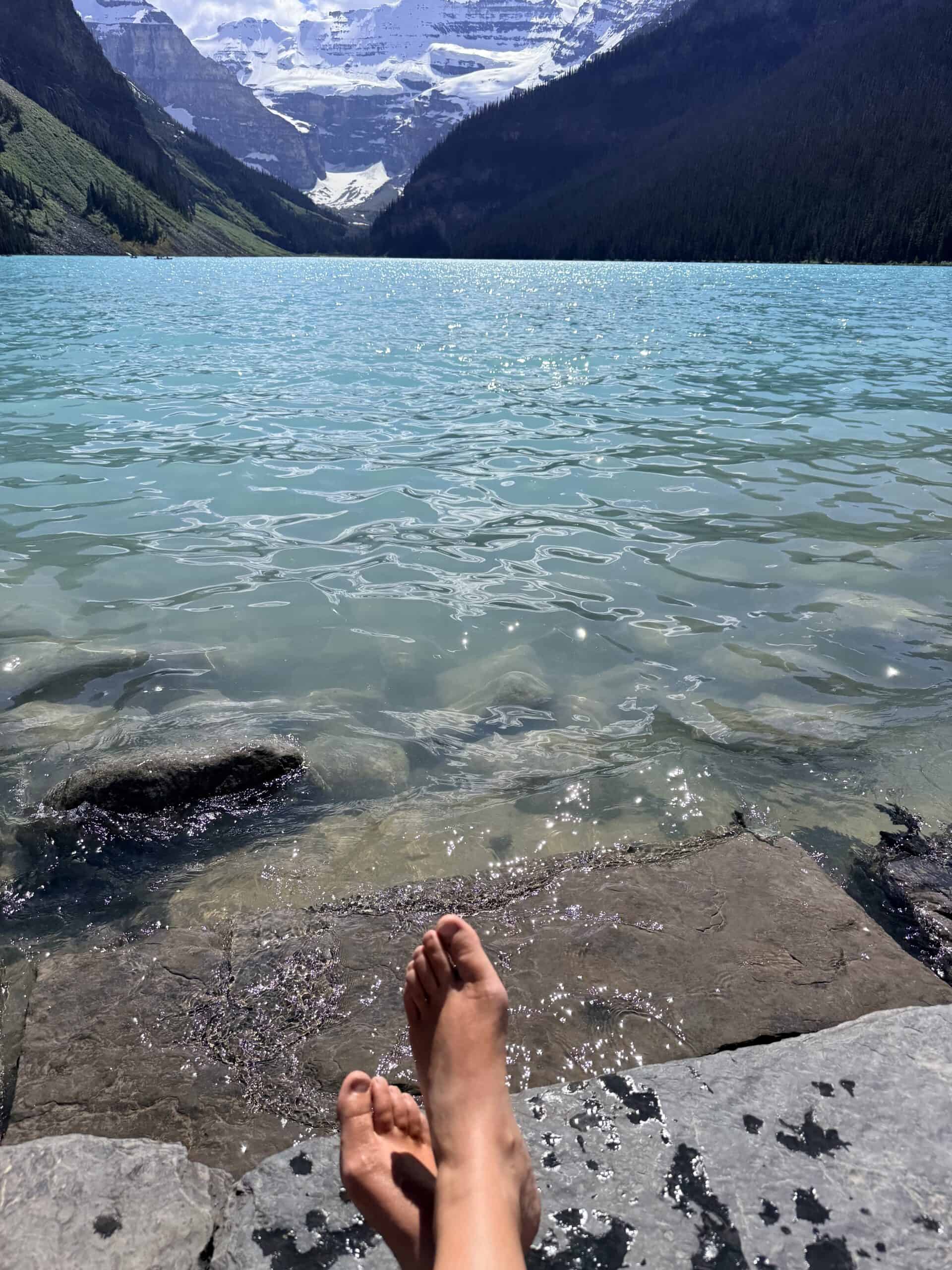 Bare feet resting on sunlit shoreline rocks overlooking Lake Louise and snow-covered mountains.