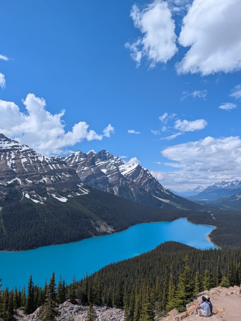 Turquoise Peyto Lake surrounded by evergreen forest and snow-streaked mountains, with two visitors sitting on rocks overlooking the lake.