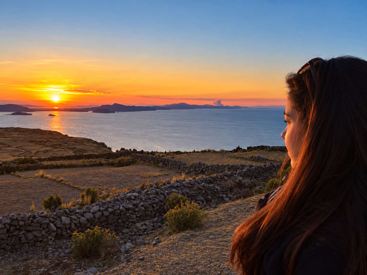 Sunset over Lake Titicaca from Pachamama in Peru, with rocky terraces in the foreground and a woman looking toward the horizon.