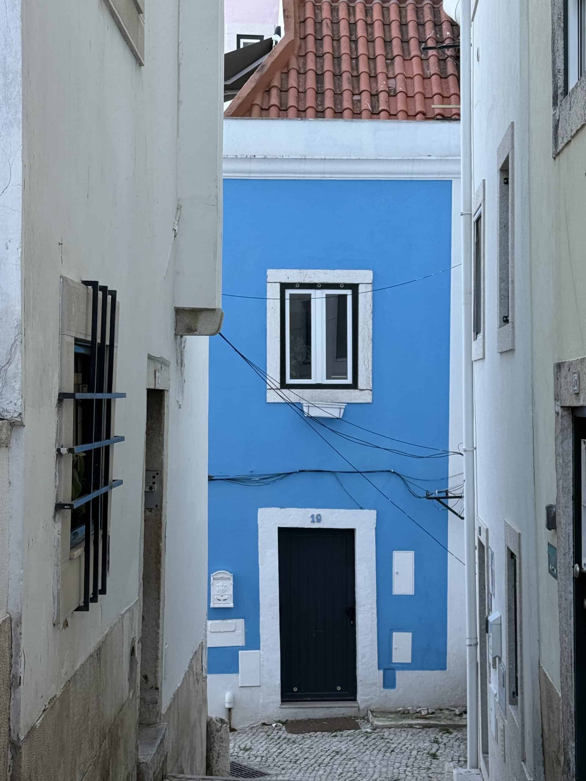 Bright blue house with white window frames, a black door and a terracotta roof at the end of a narrow cobblestone street in Alfama, Lisbon.