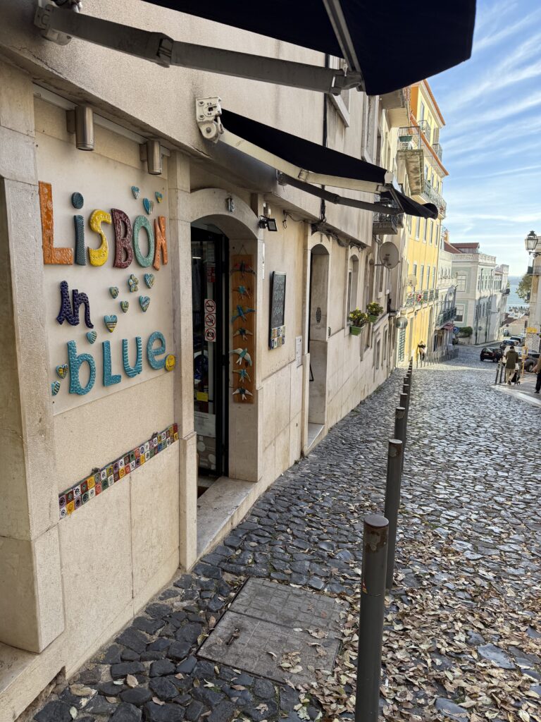 Colourful Lisbon in Blue sign beside a narrow cobblestone street descending toward the Tagus River.