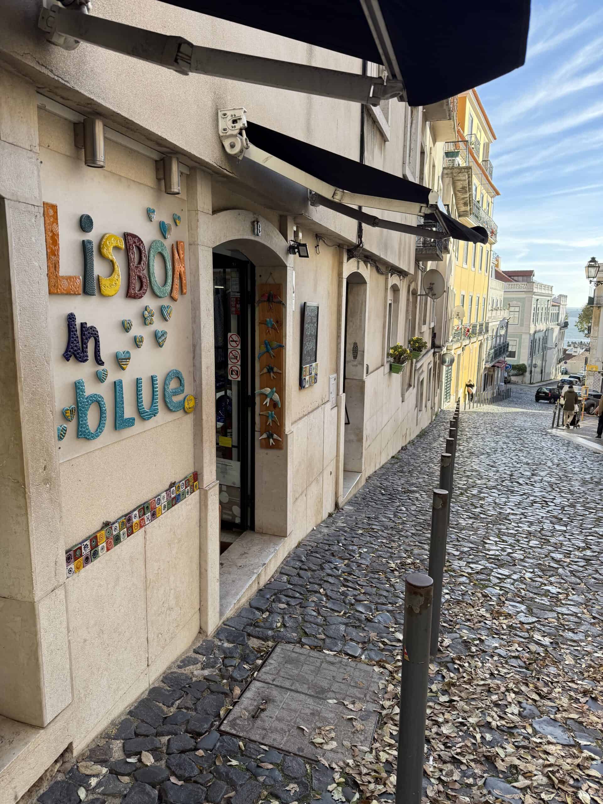 Colourful Lisbon in Blue sign beside a narrow cobblestone street descending toward the Tagus River in Alfama.