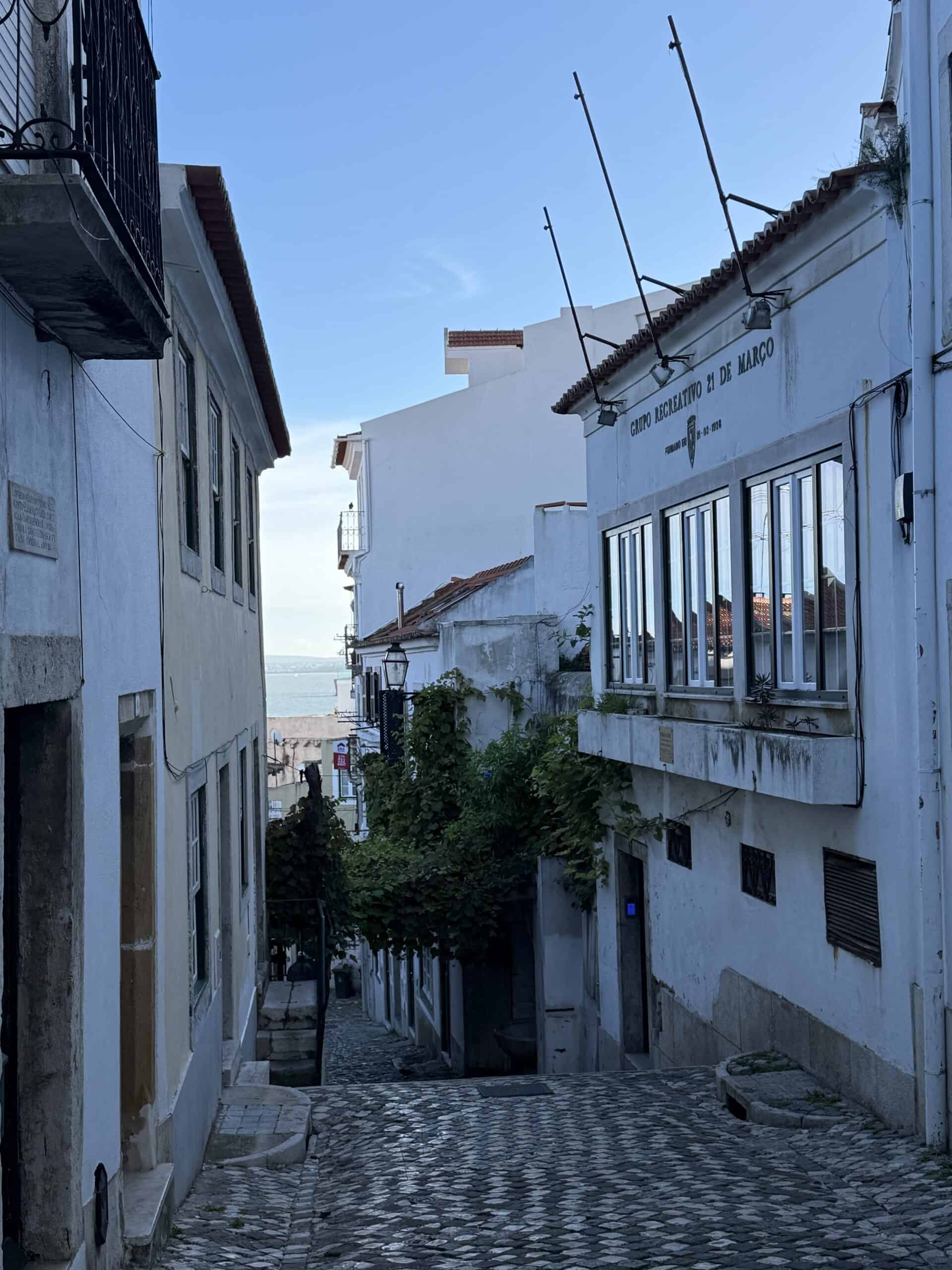 Narrow cobblestone street between white buildings with greenery and a glimpse of the Tagus River in Alfama.