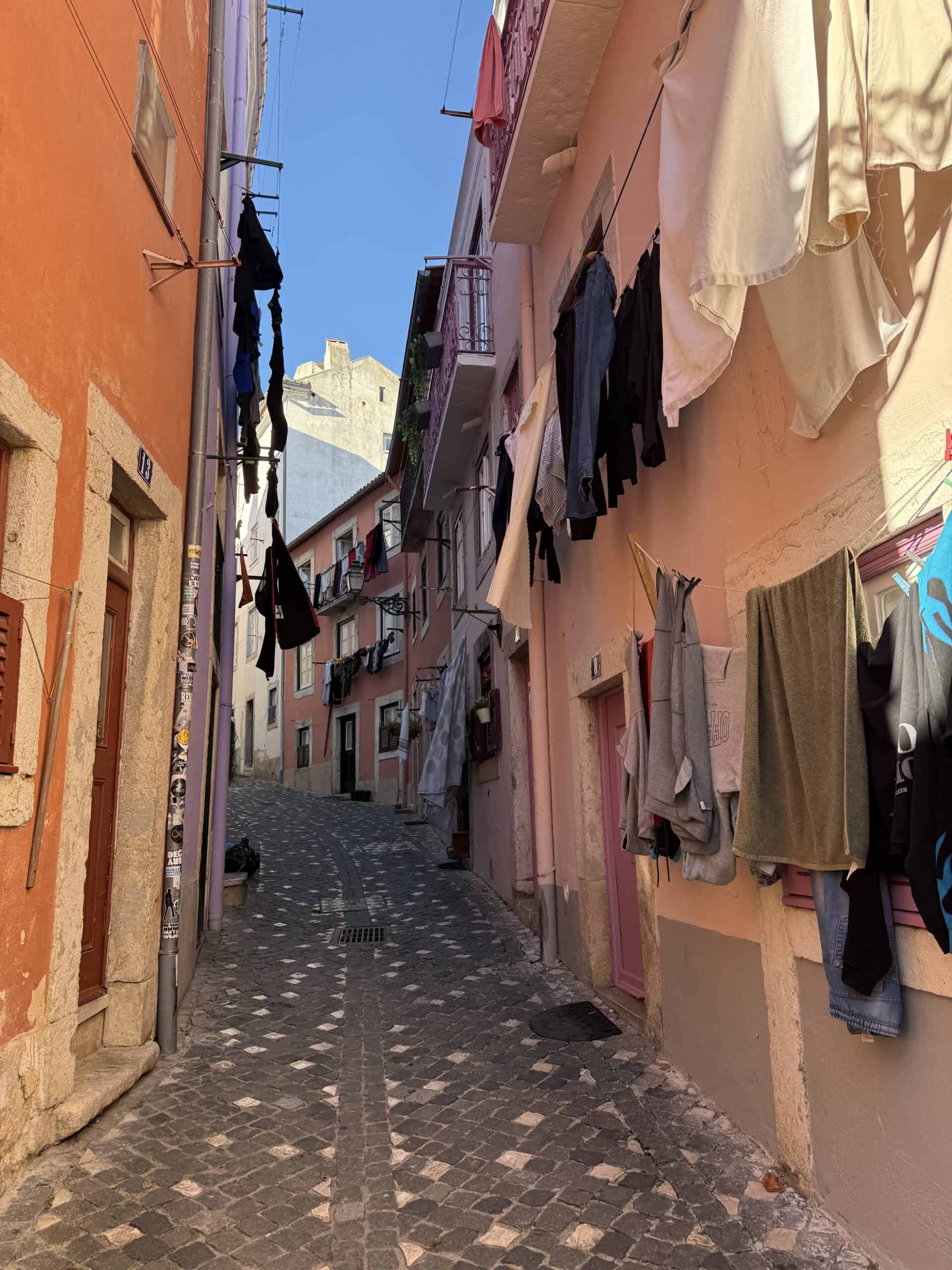 Narrow cobblestone alley in Alfama, Lisbon, lined with pink and orange buildings and laundry hanging overhead.