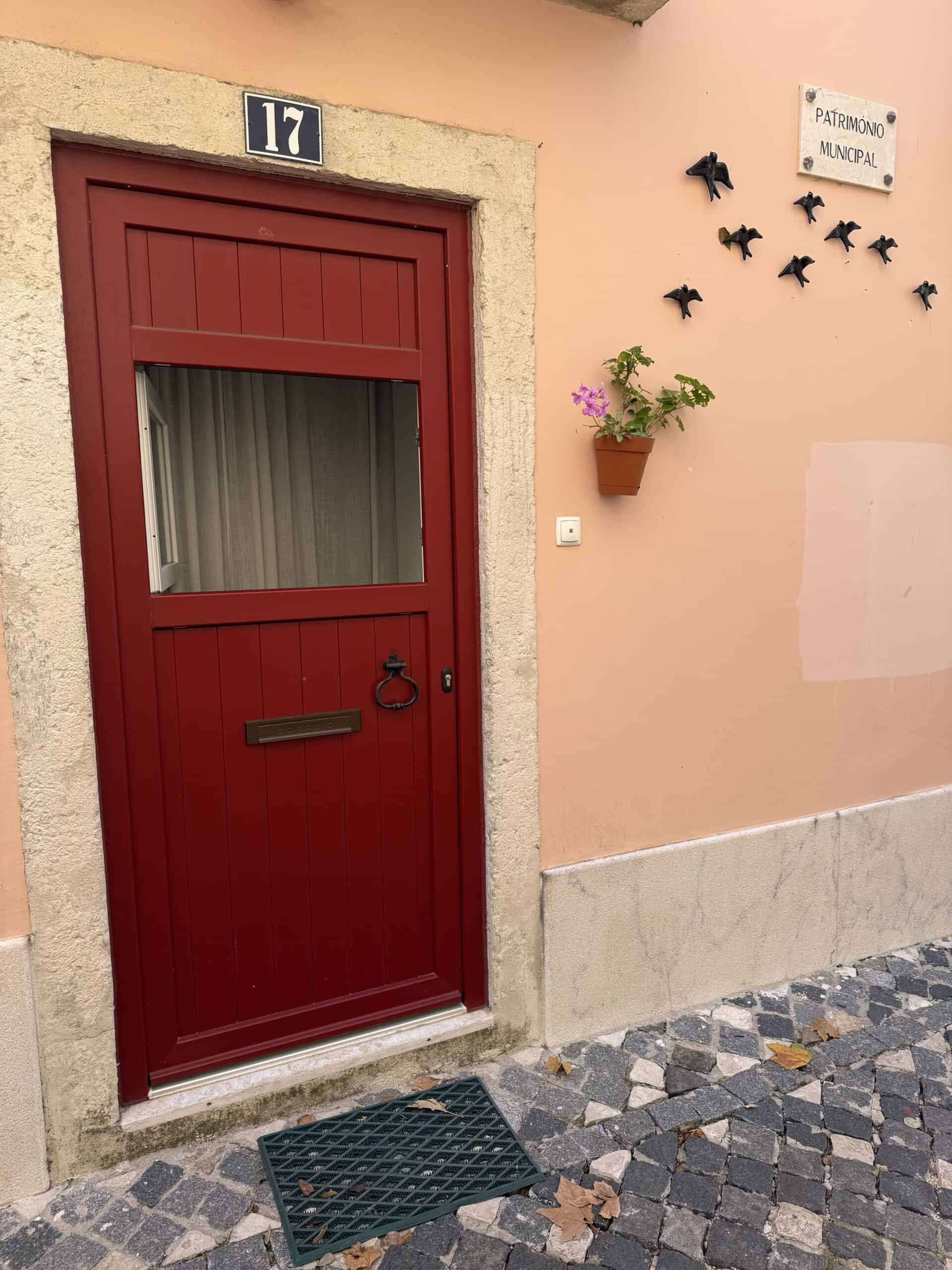 Traditional red wooden door on a peach-coloured building with decorative birds and a small flowerpot in Alfama, Lisbon.