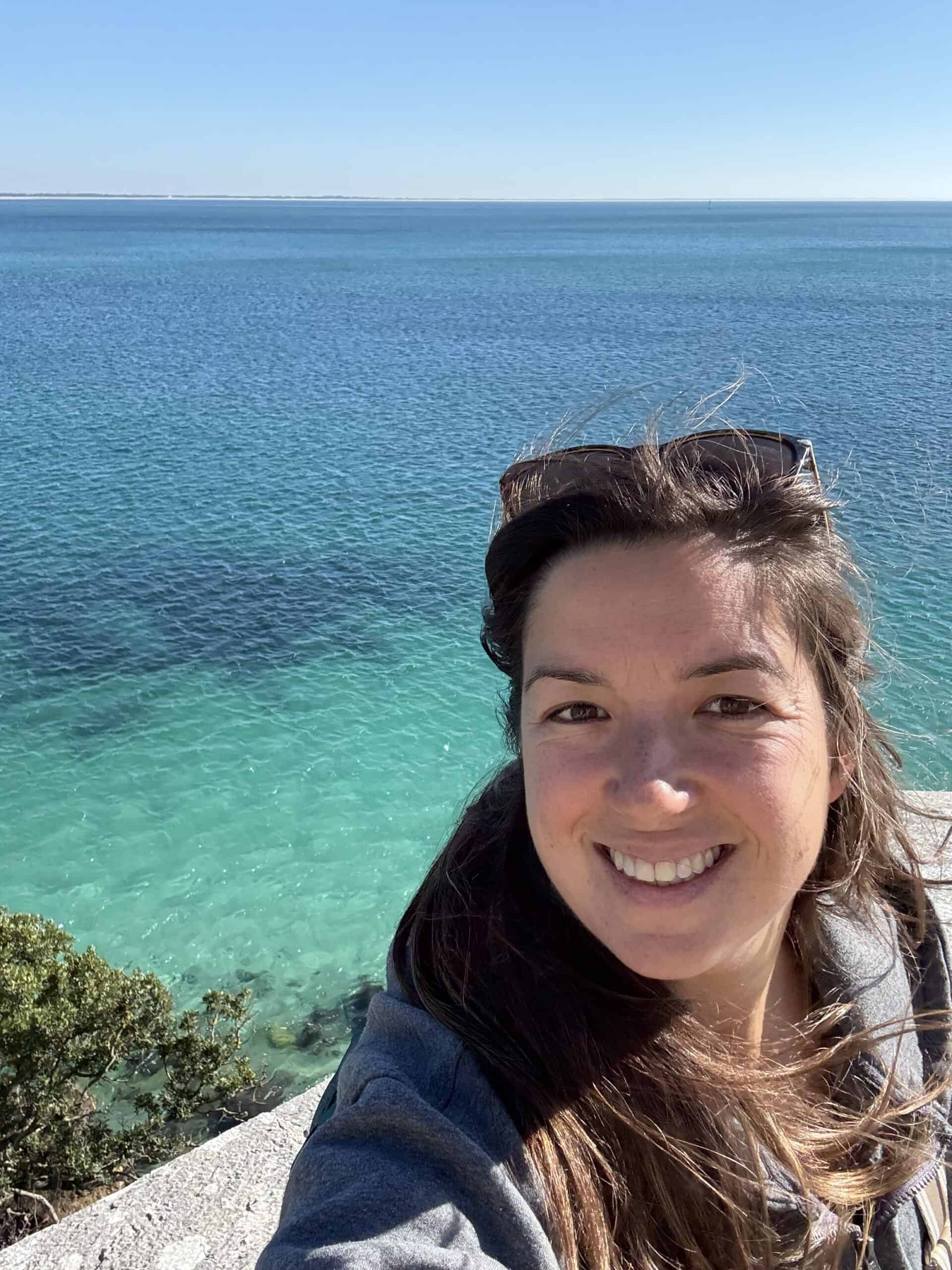 Annie smiling with the crystal-clear turquoise waters of Arrábida Natural Park, Portugal, in the background