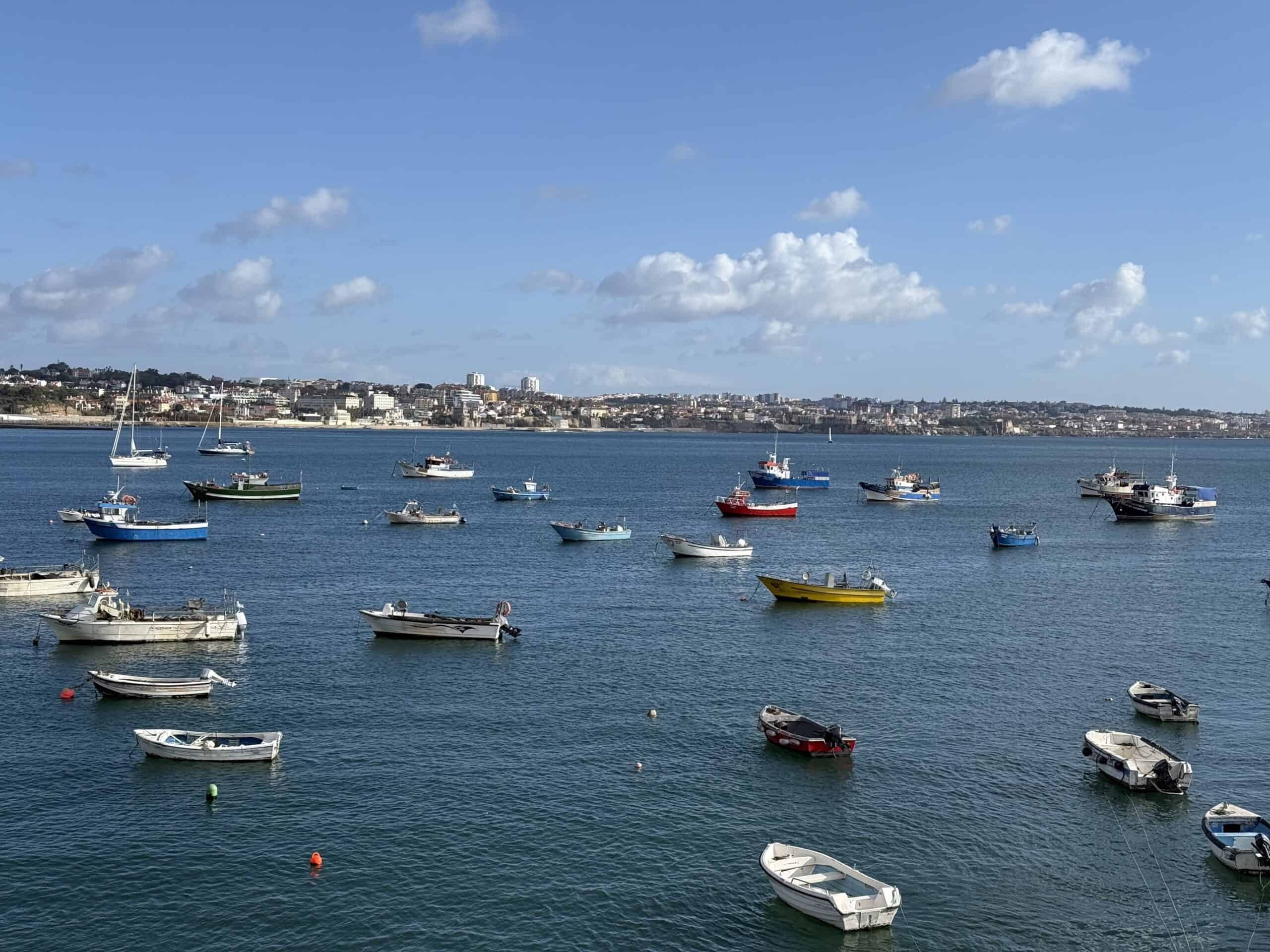 Colourful fishing boats anchored in the calm blue waters of Cascais Bay, Portugal, with the coastal town in the background.