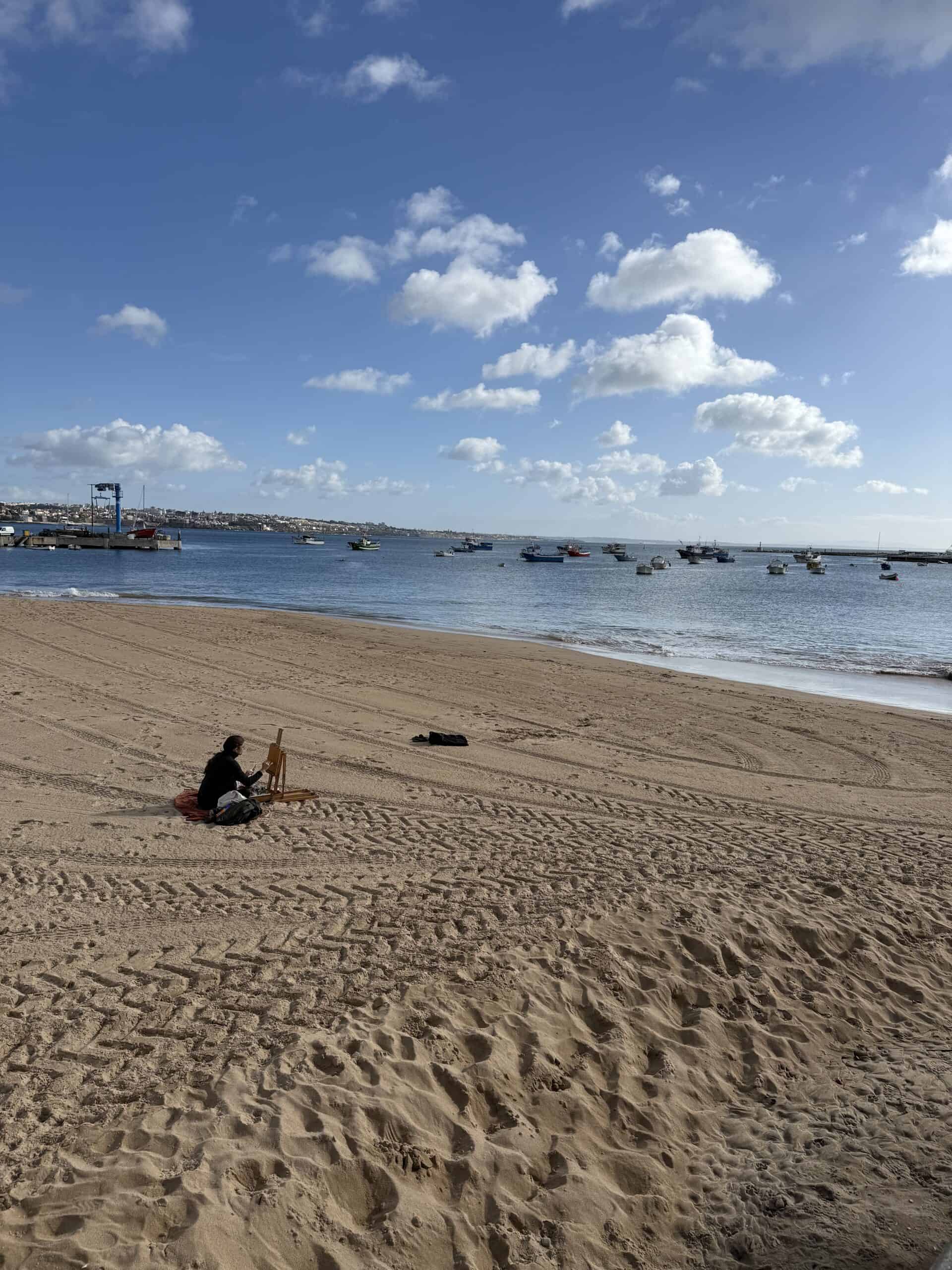 Artist painting beside the beach with fishing boats in the bay at Cascais, Portugal