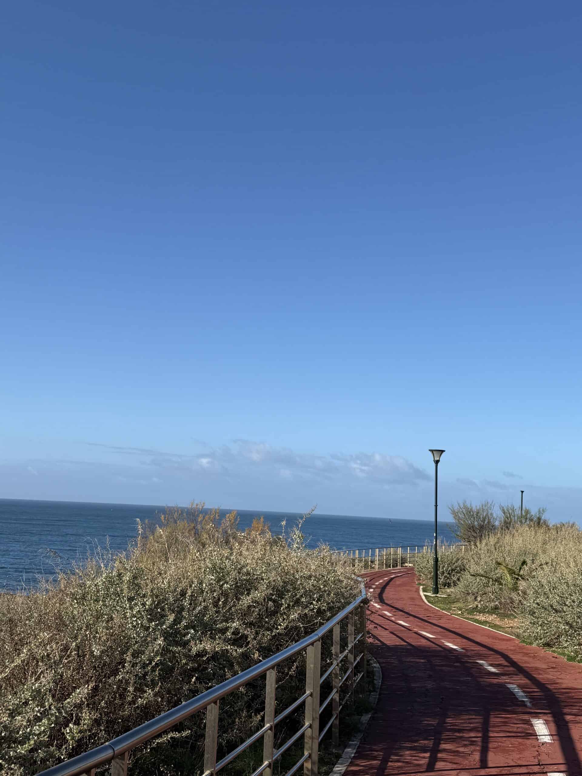Red coastal walking and cycling path curving beside the Atlantic Ocean in Cascais, Portugal, with railings and coastal vegetation.