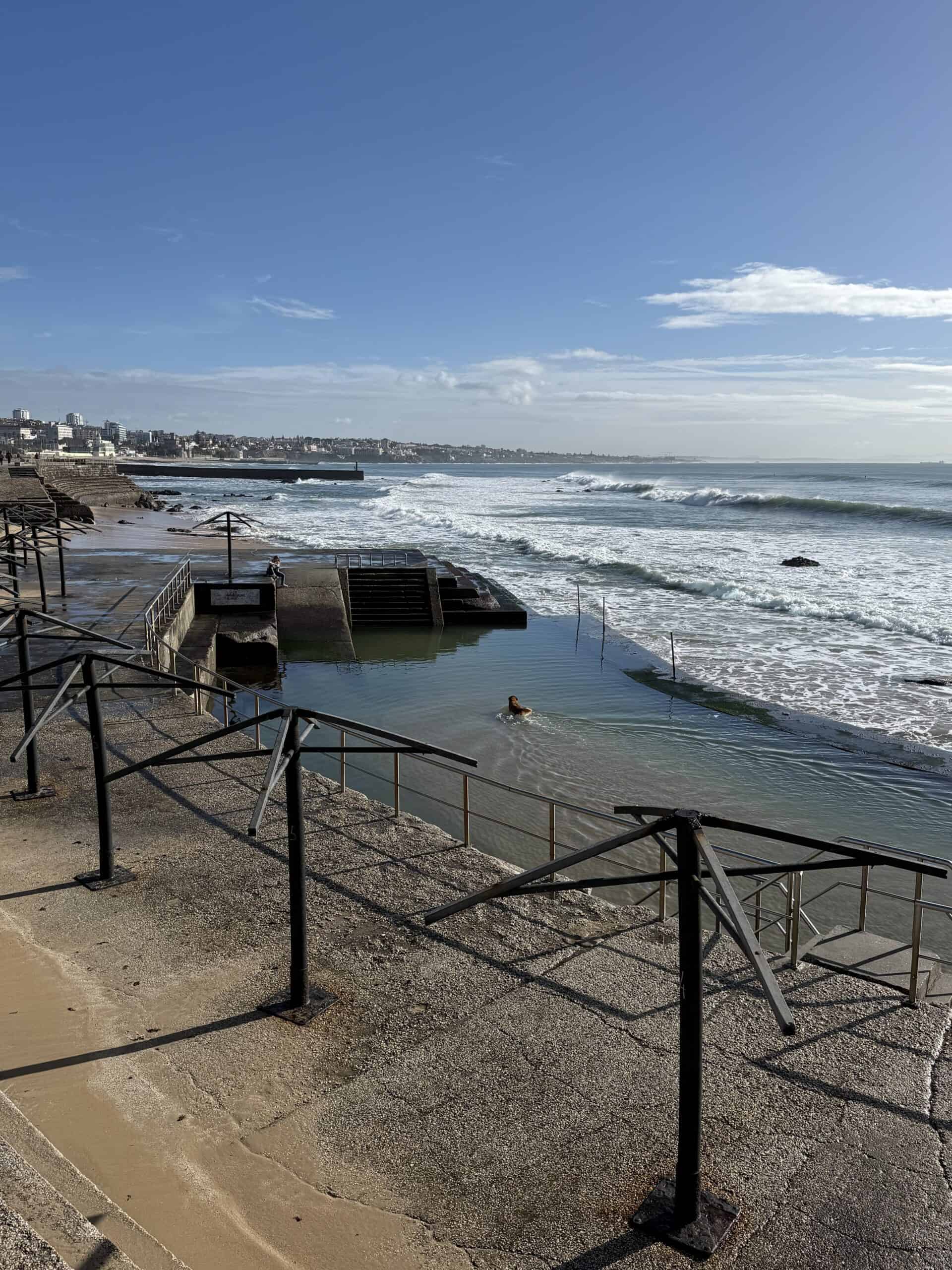 Oceanfront tidal swimming pool along the Cascais coastline in Portugal, with concrete steps, railings and Atlantic waves breaking nearby.