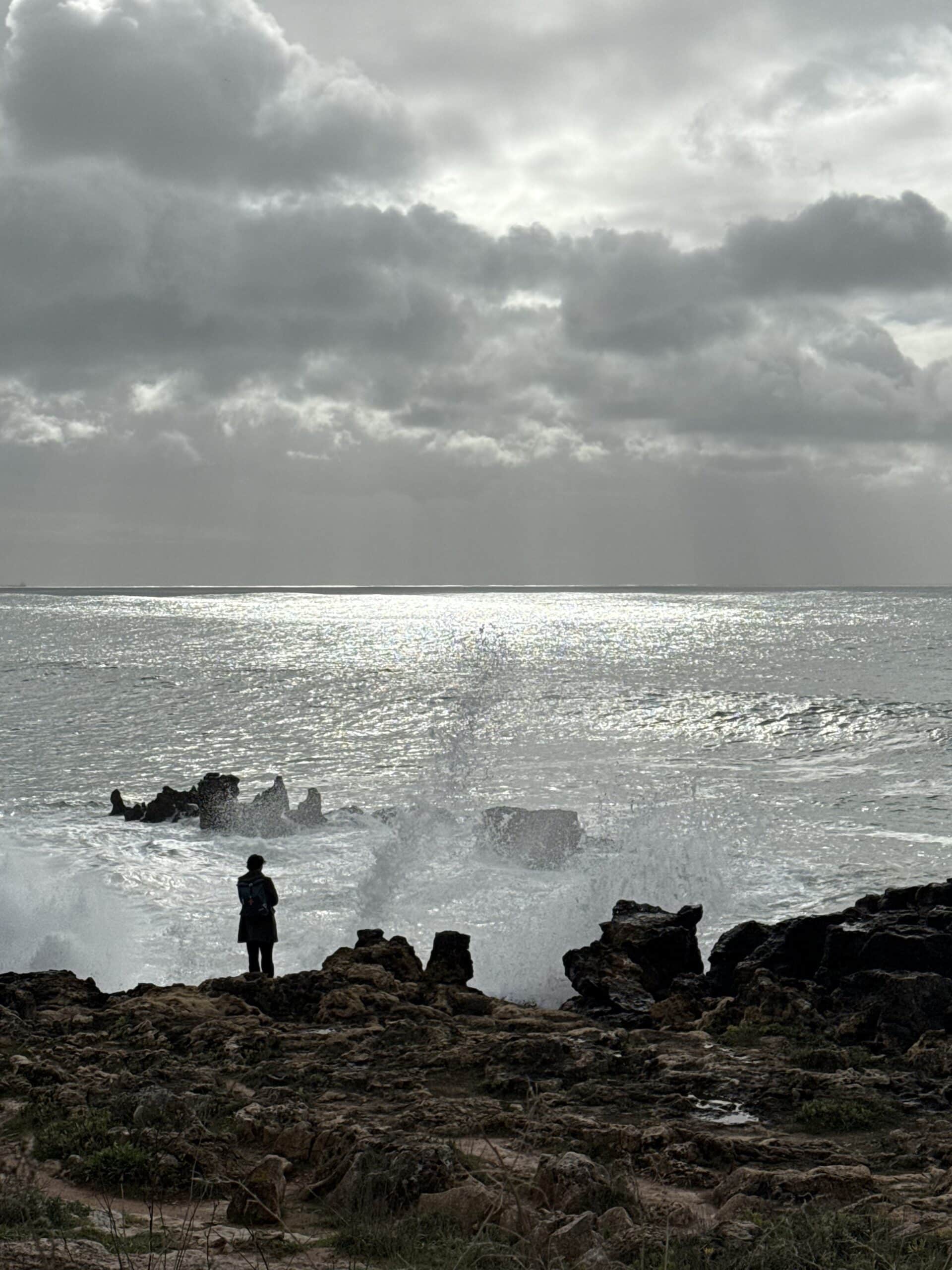 Person standing on the rocky coastline of Cascais as a powerful Atlantic wave sends a tall plume of sea spray into the air.