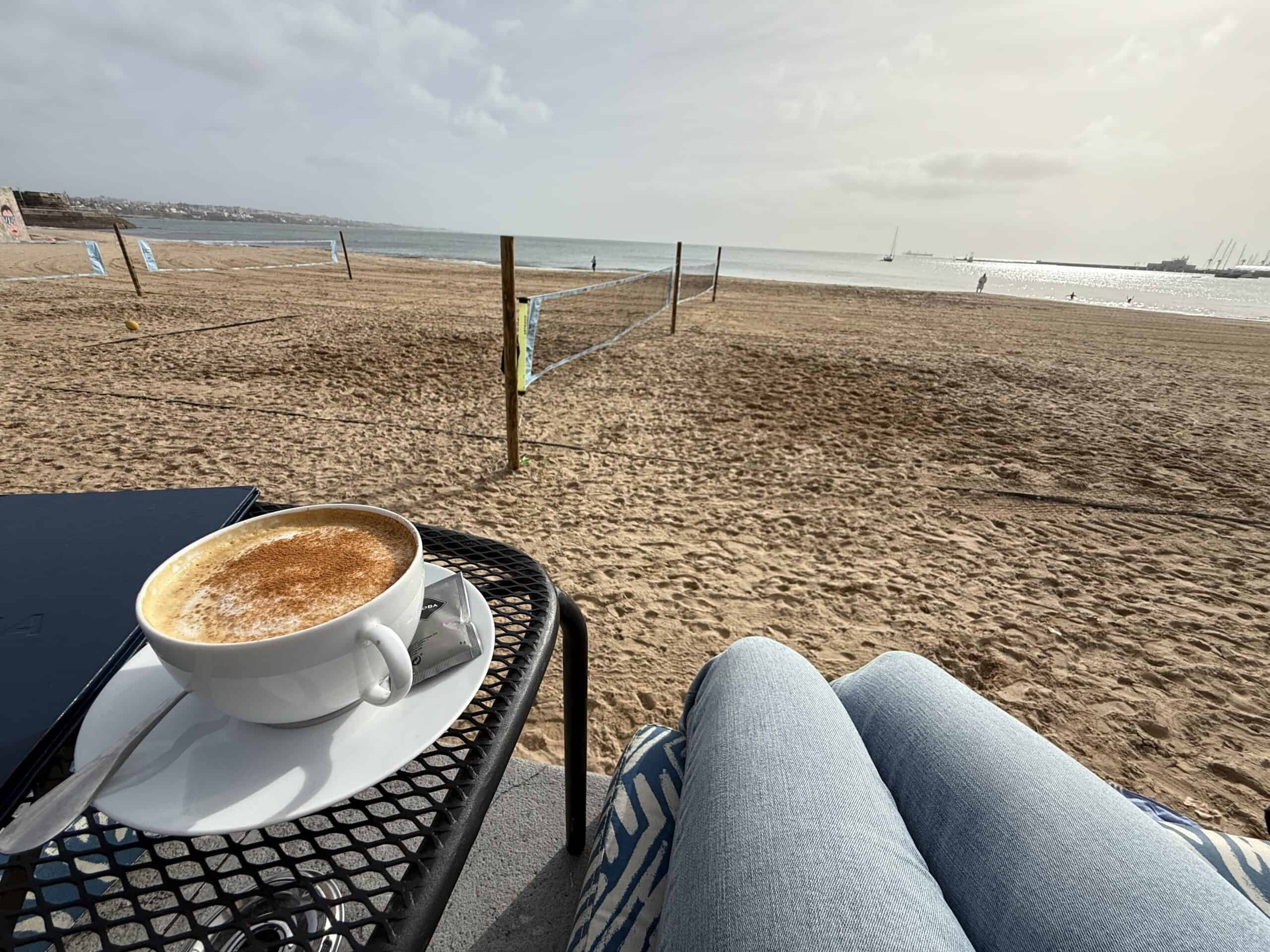 Cappuccino on an outdoor table at Emma's Café in Cascais, overlooking the sandy beach, volleyball courts and Atlantic Ocean.