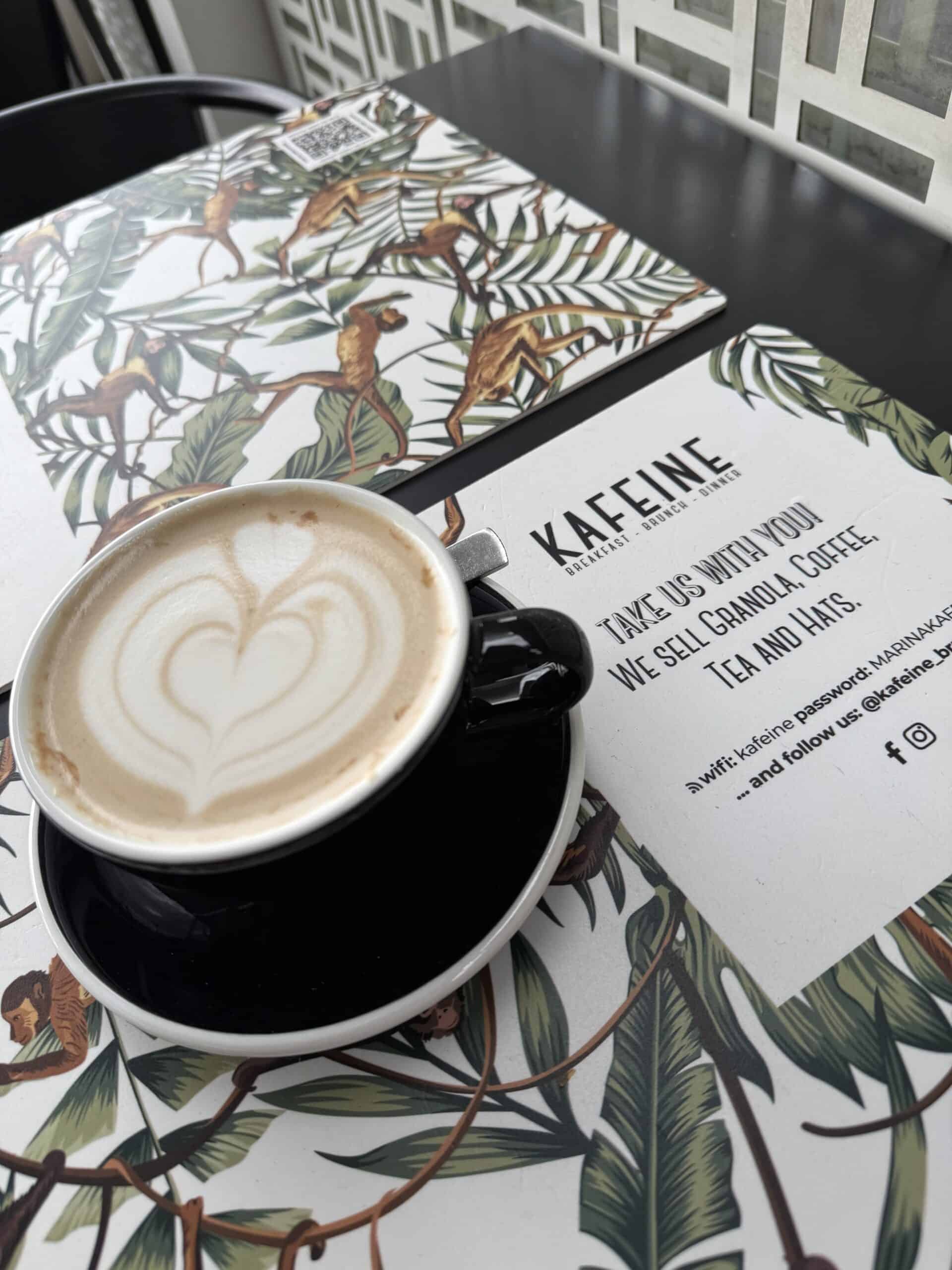 Cappuccino with heart-shaped latte art at Kafeine café in Cascais Marina, beside a tropical-patterned menu.