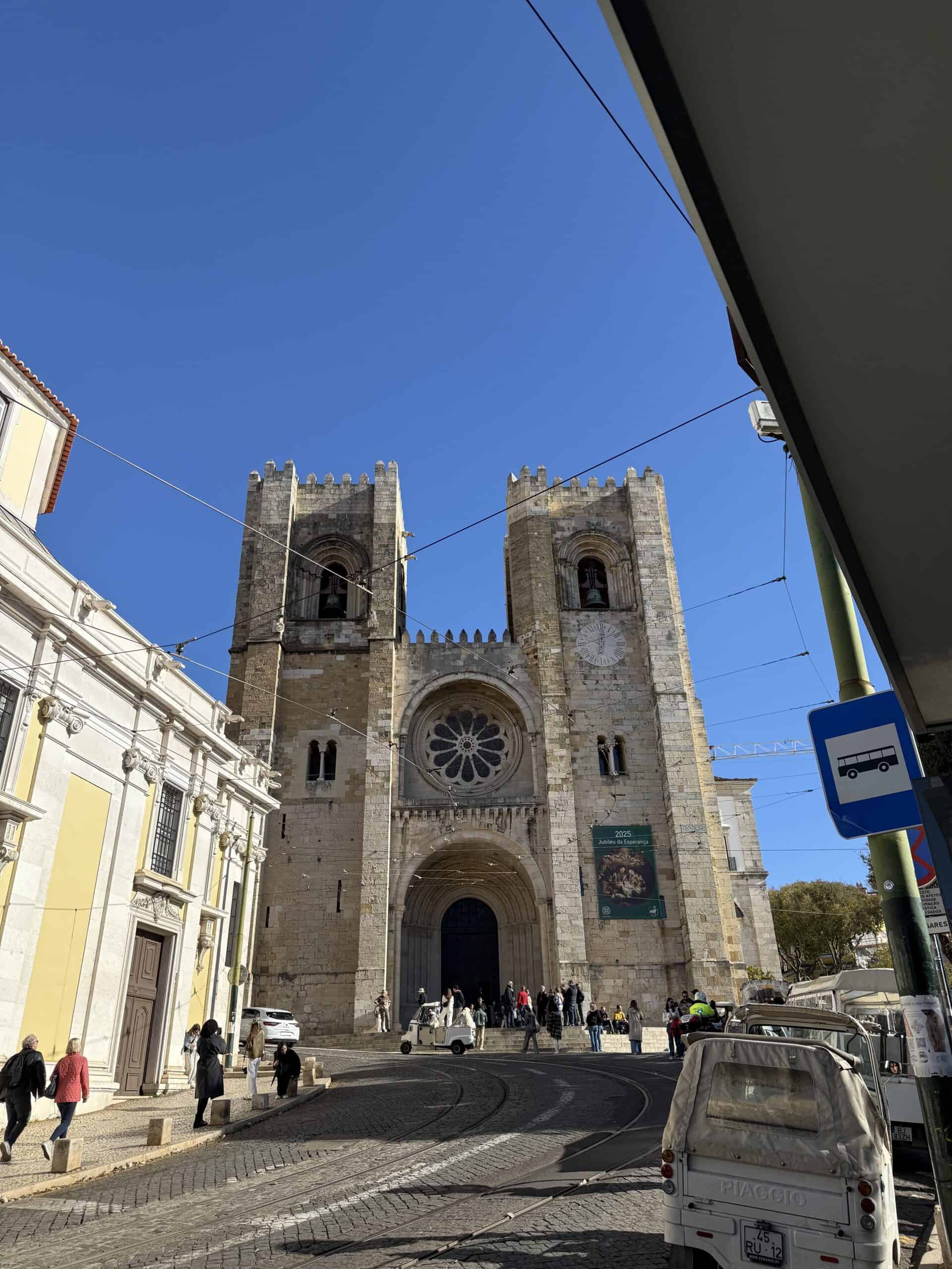 Historic façade of Lisbon Cathedral (Sé de Lisboa) with twin stone towers, a rose window and tram tracks along the cobblestone street in Alfama, Portugal.