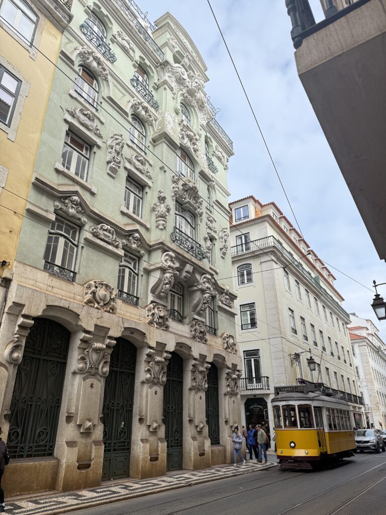 Traditional yellow Lisbon tram passing an ornate historic building with decorative stonework and wrought-iron balconies.