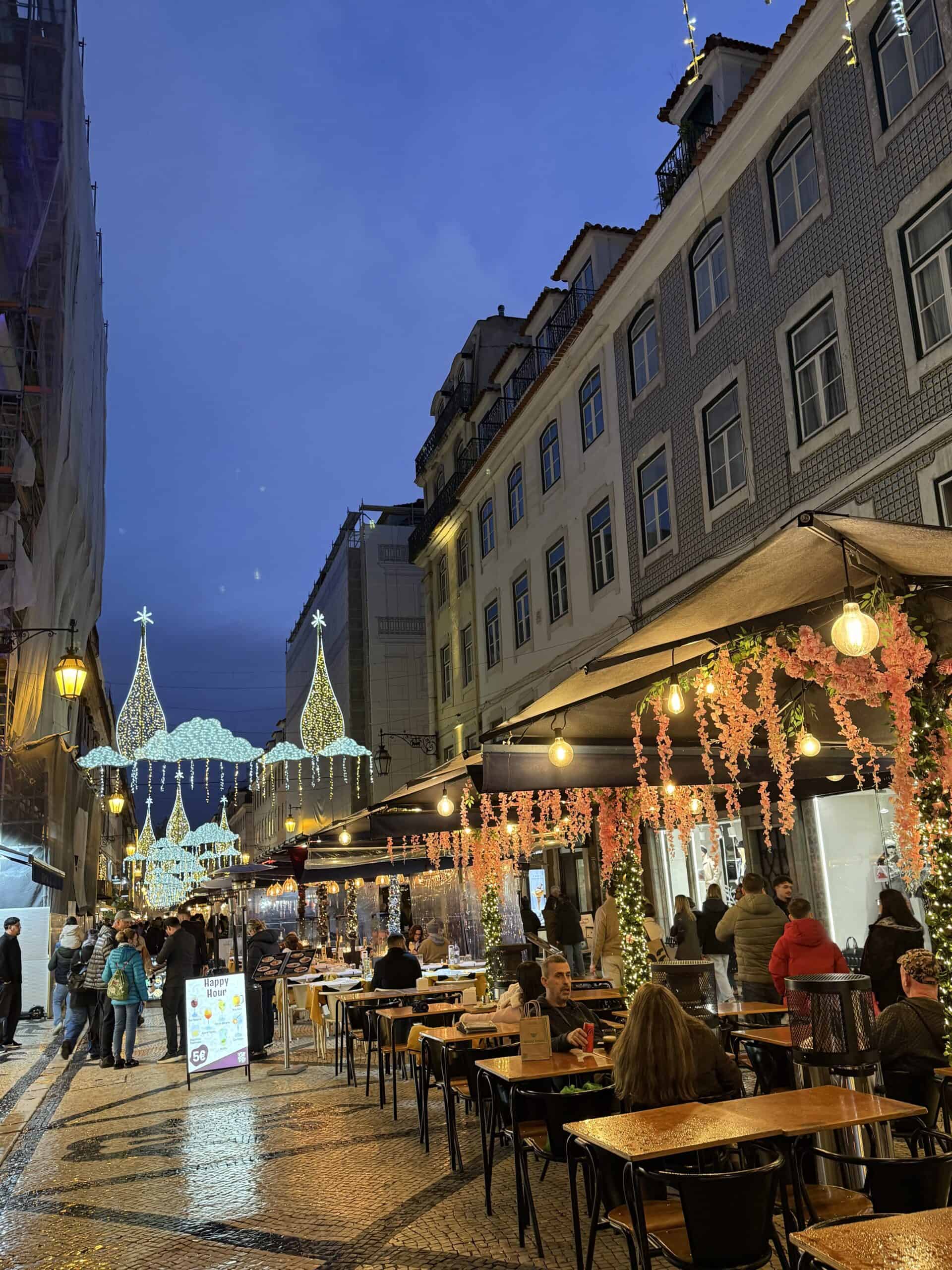 Outdoor restaurant terraces along a cobblestone street in Lisbon decorated with Christmas lights and hanging flowers.