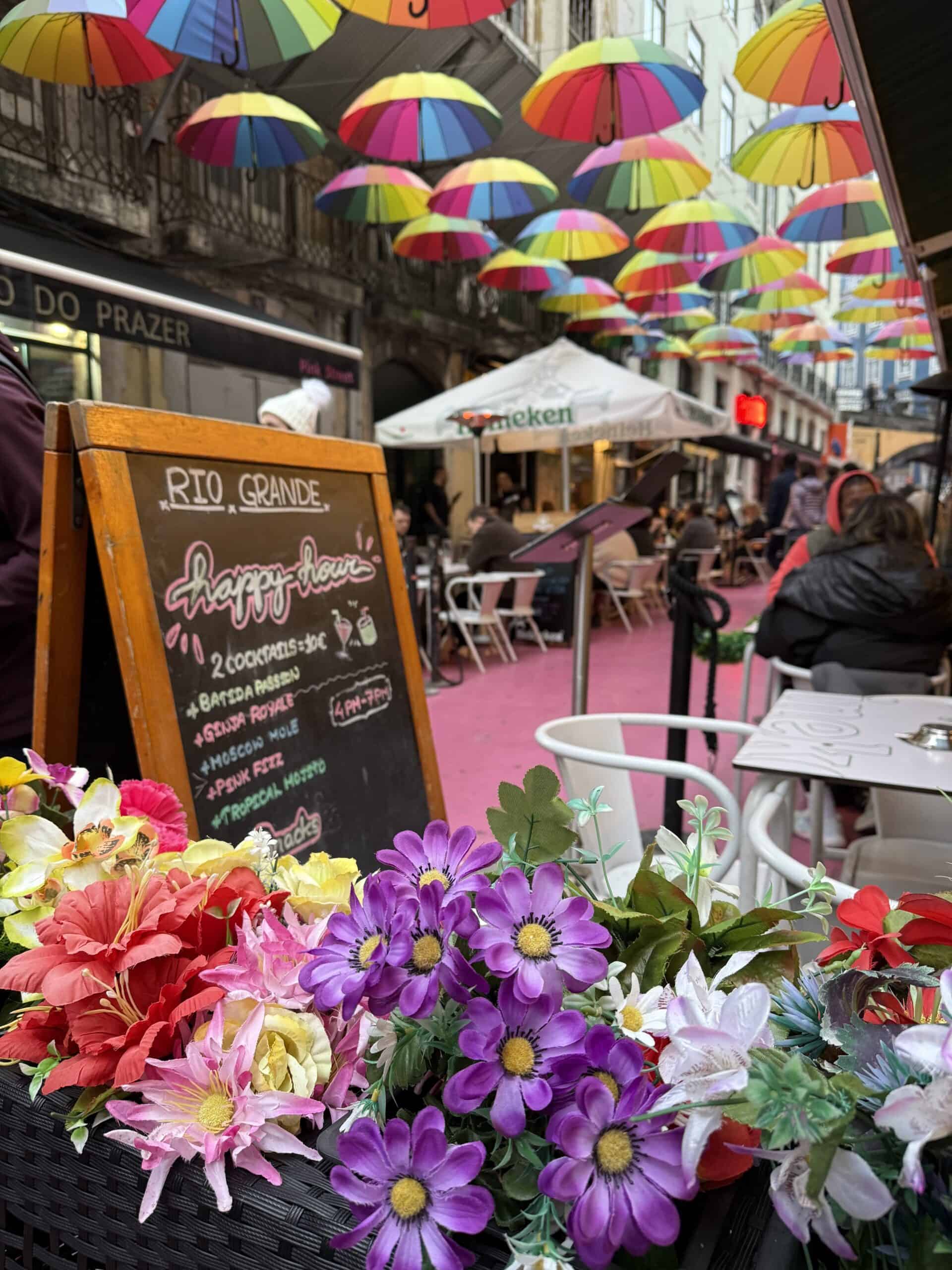Colourful rainbow umbrellas and flowers decorating Pink Street in Lisbon, with outdoor café terraces.