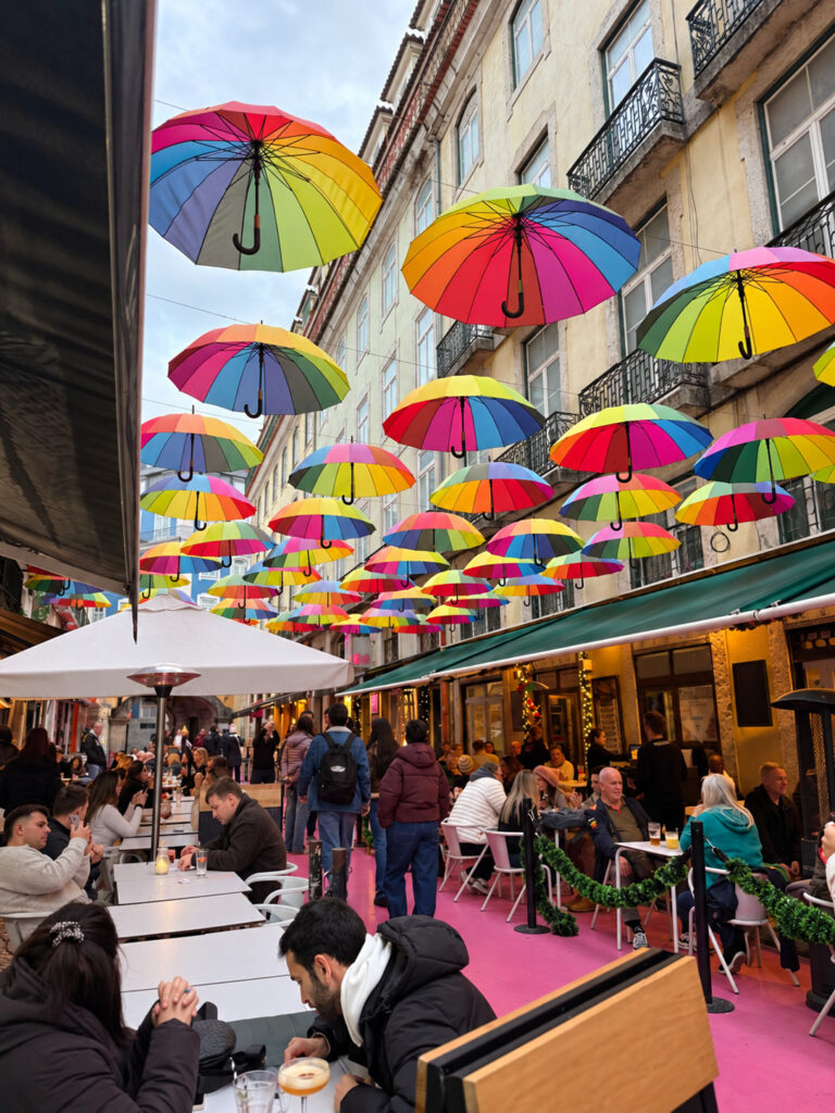 Colourful rainbow umbrellas hanging above Pink Street in Lisbon, Portugal, with outdoor café terraces, people dining and a vibrant pink walkway.