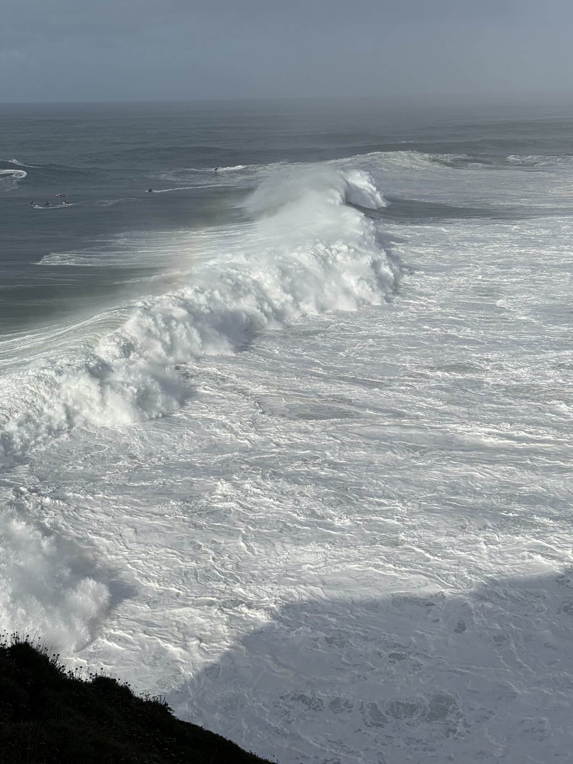 Massive breaking waves and whitewater at Praia do Norte in Nazaré, Portugal, with small boats visible offshore