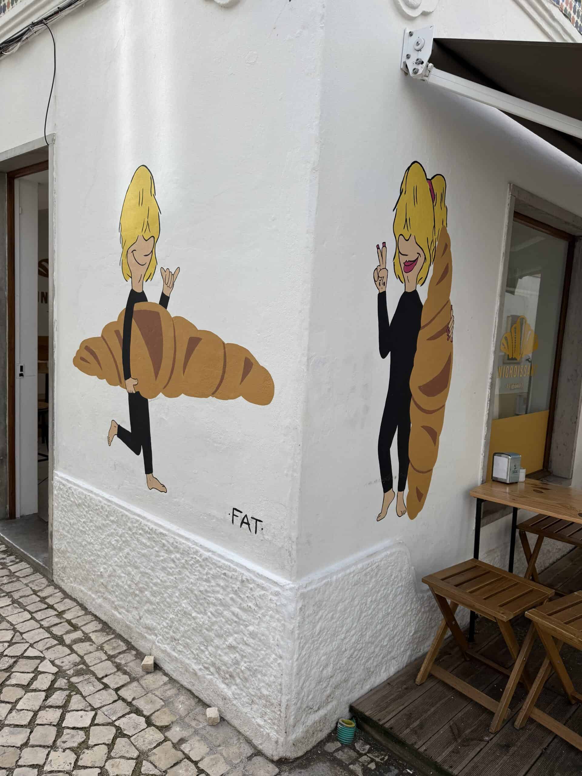 Colourful mural of two cartoon figures holding oversized croissants on the exterior walls of a bakery in Nazaré, Portugal