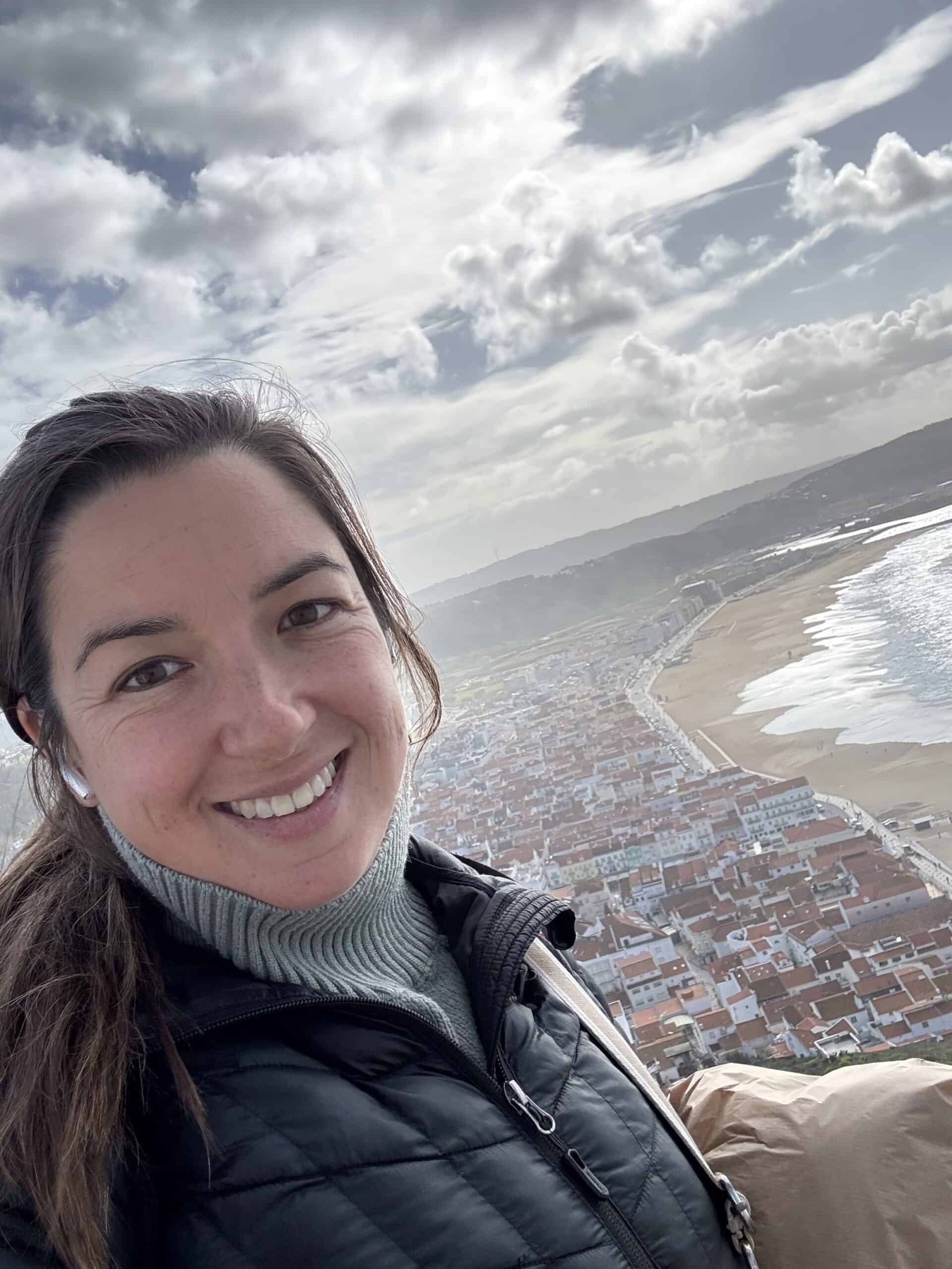 Travel blogger smiling from a viewpoint overlooking Nazaré's red-roofed town and Praia da Nazaré beach in Portugal