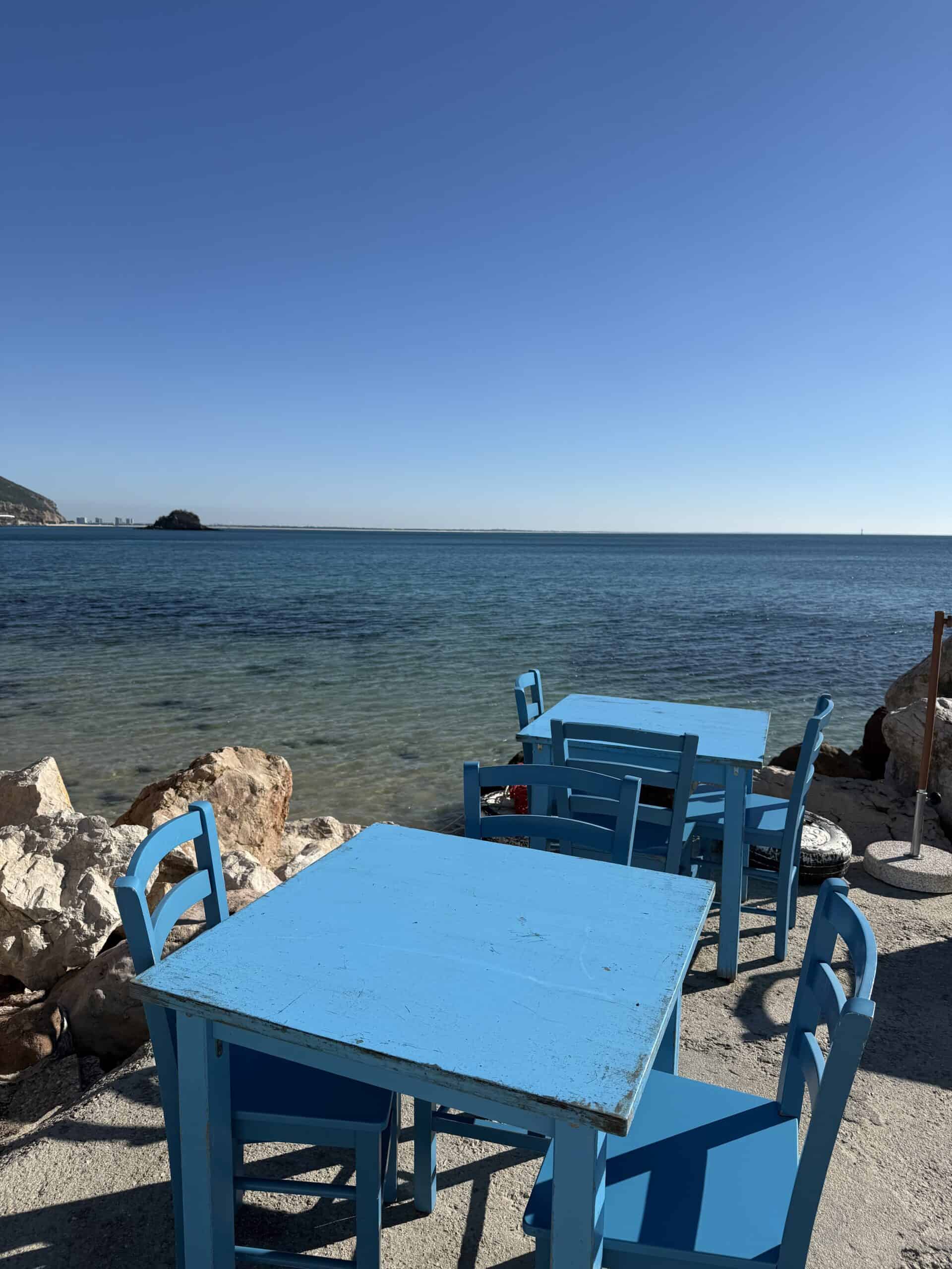 Bright blue restaurant tables and chairs overlooking the crystal-clear waters of Portinho da Arrábida, Portugal