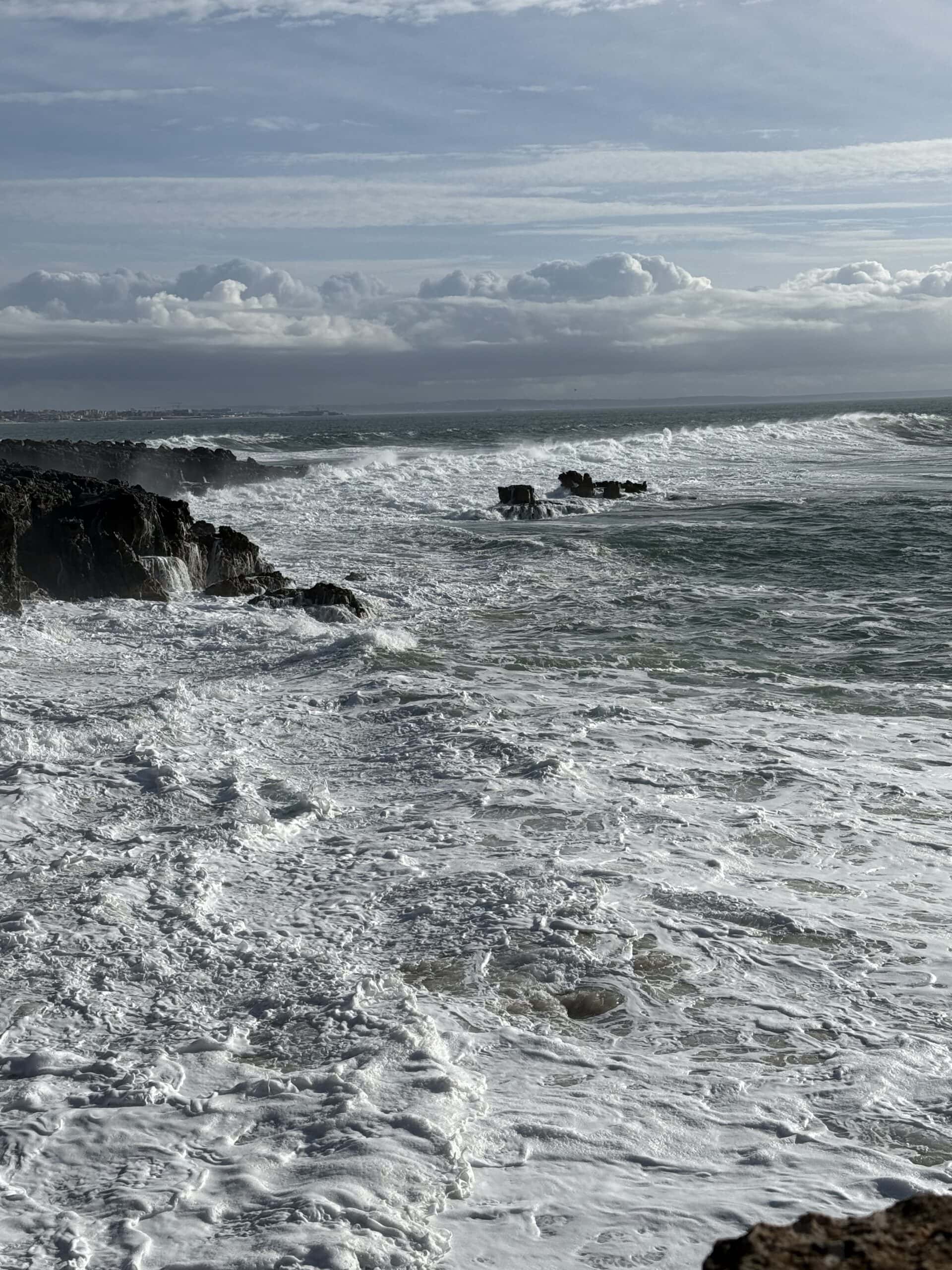 Powerful Atlantic waves breaking against dark coastal rocks in Cascais, Portugal, with white sea foam covering the water.