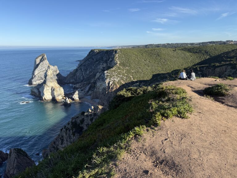 Two visitors sitting at a viewpoint overlooking Praia da Ursa’s towering rock formations and turquoise water.