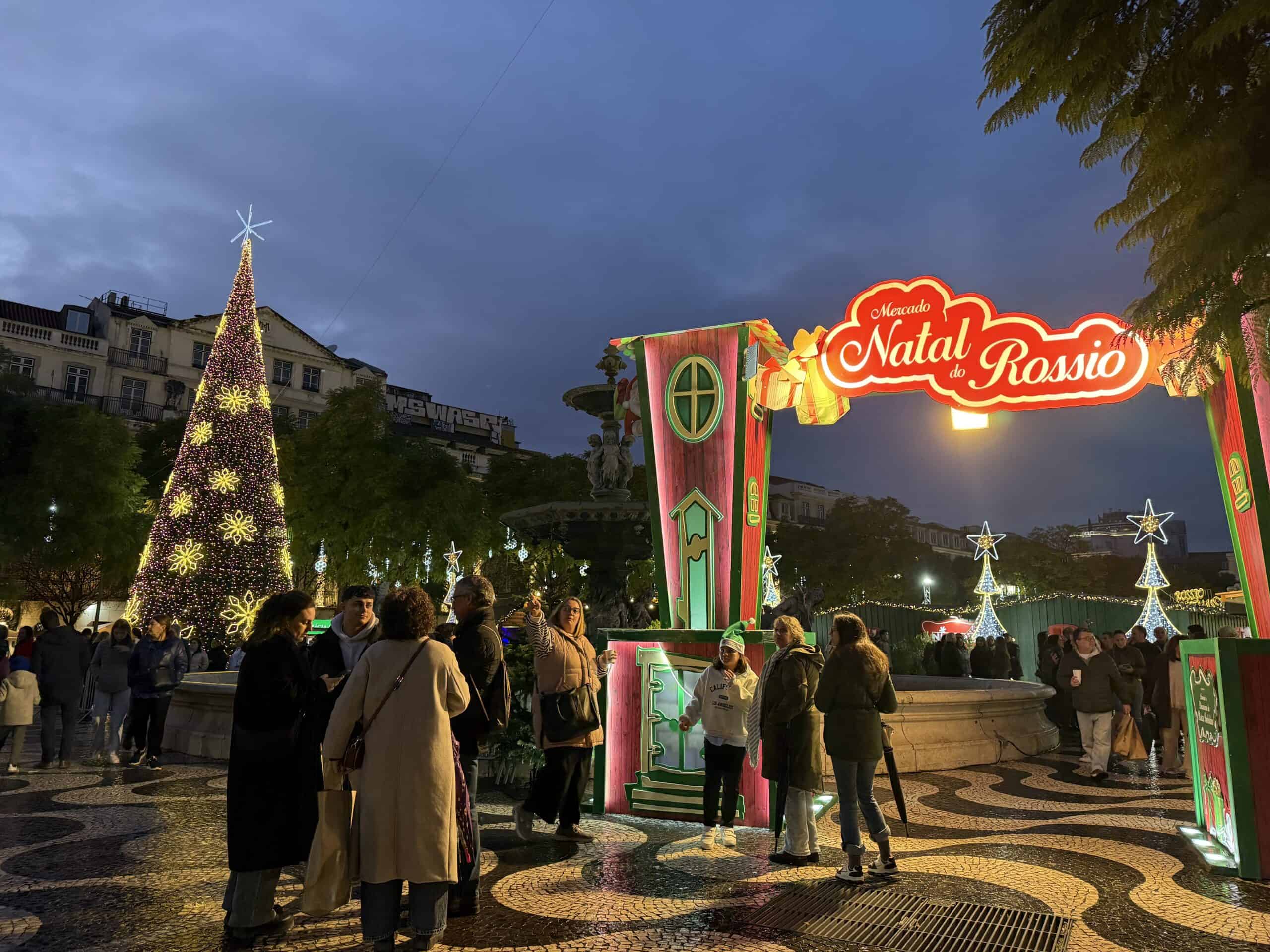 Illuminated entrance to the Mercado de Natal do Rossio Christmas market in Lisbon, with festive lights, a Christmas tree and visitors.
