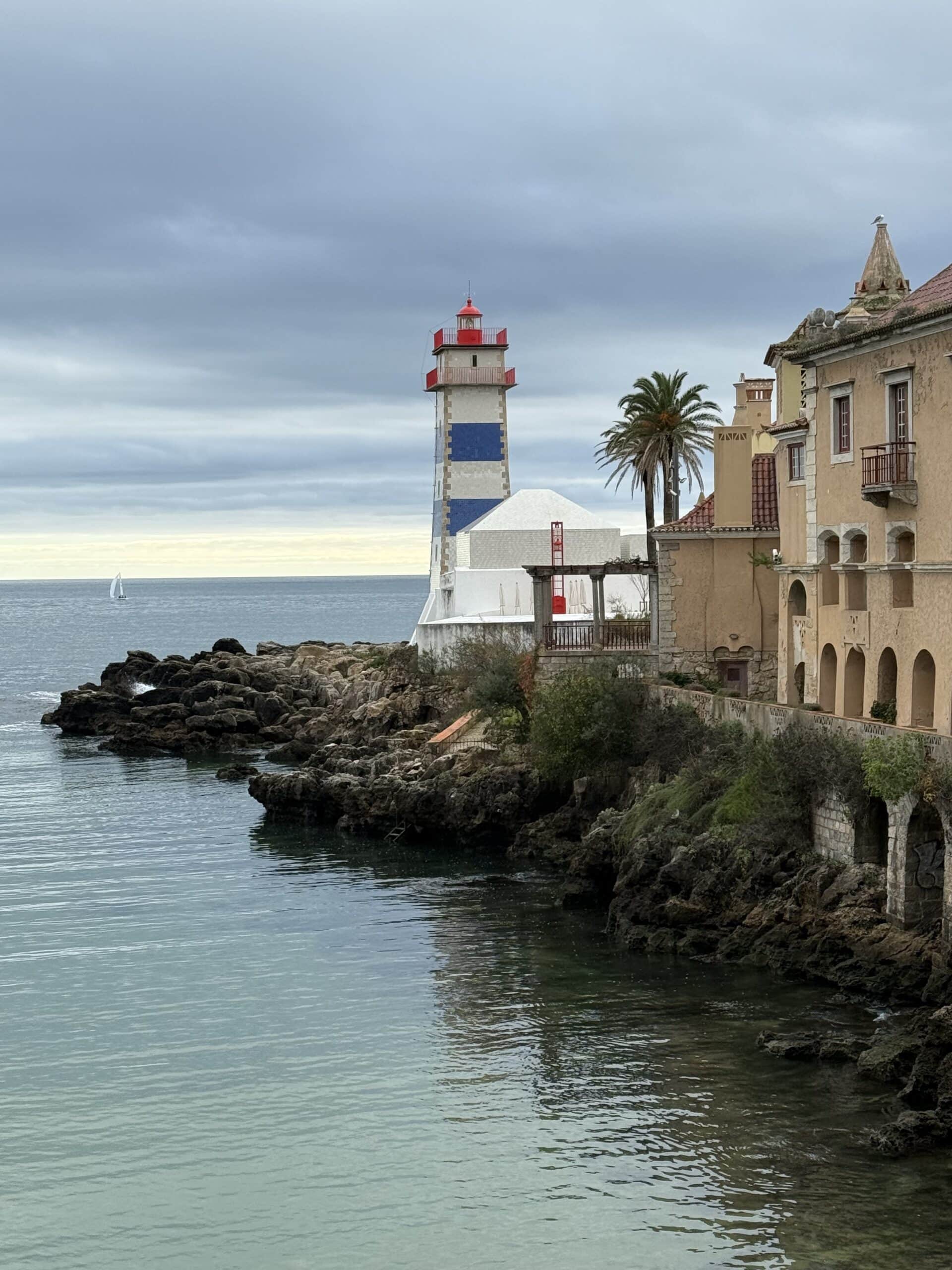 Blue-and-white Santa Marta Lighthouse beside Casa de Santa Maria on the rocky Cascais coastline under a cloudy sky.