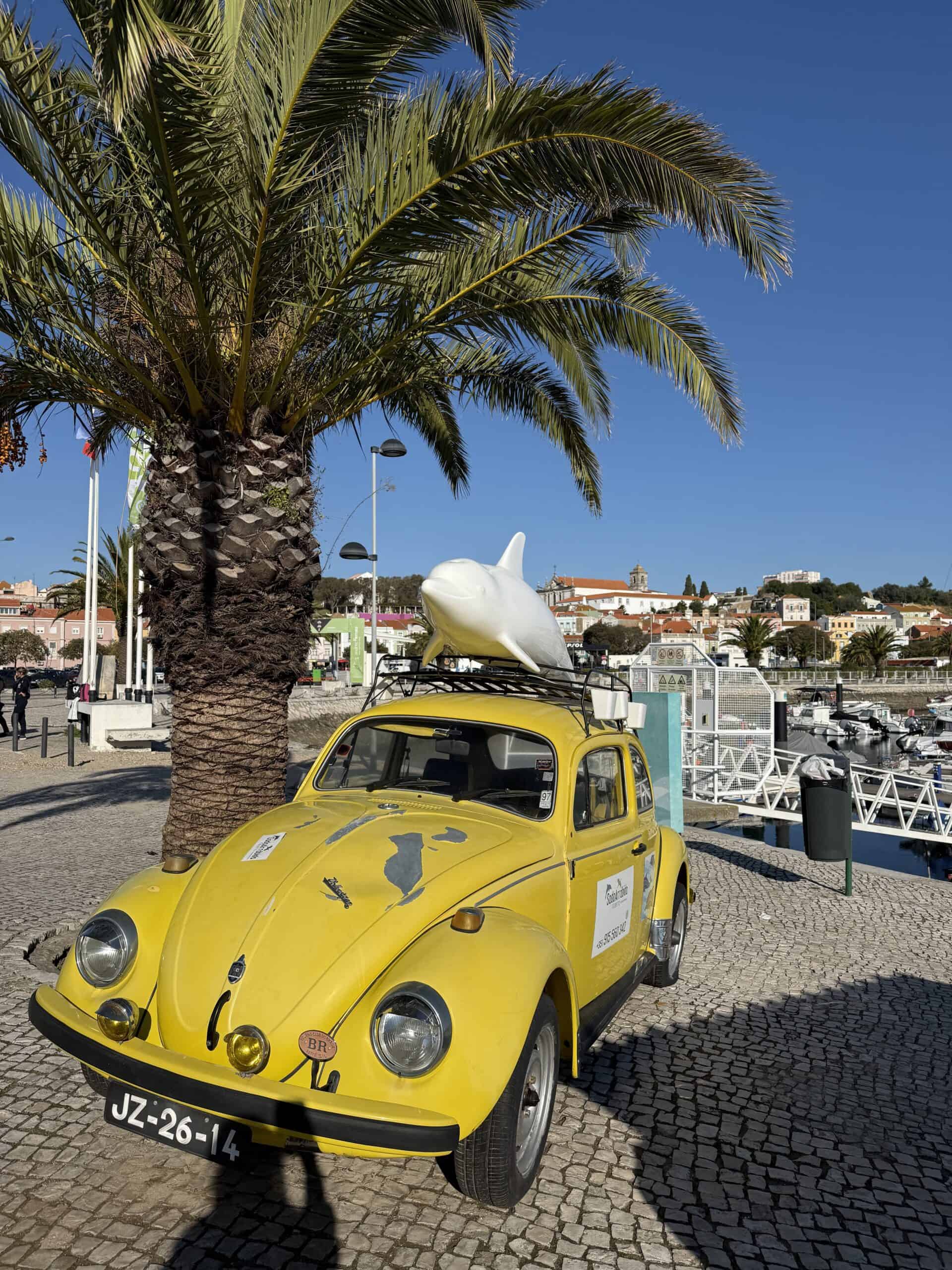 Yellow vintage Volkswagen Beetle with a dolphin sculpture on its roof beside Setúbal Marina, Portugal