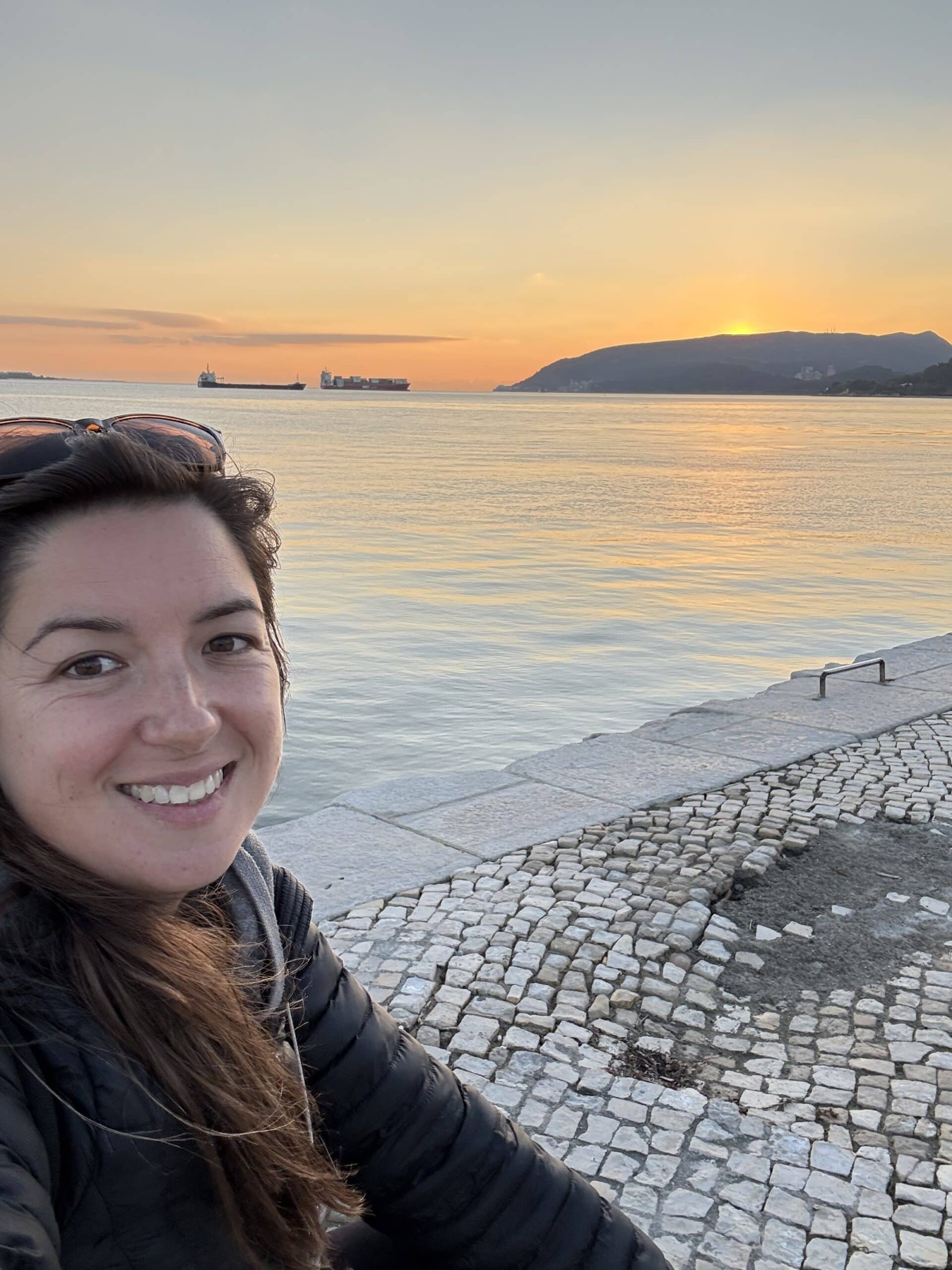 Annie smiling along Setúbal's waterfront at sunset, with golden reflections on the Sado Estuary and the Arrábida mountains in the background