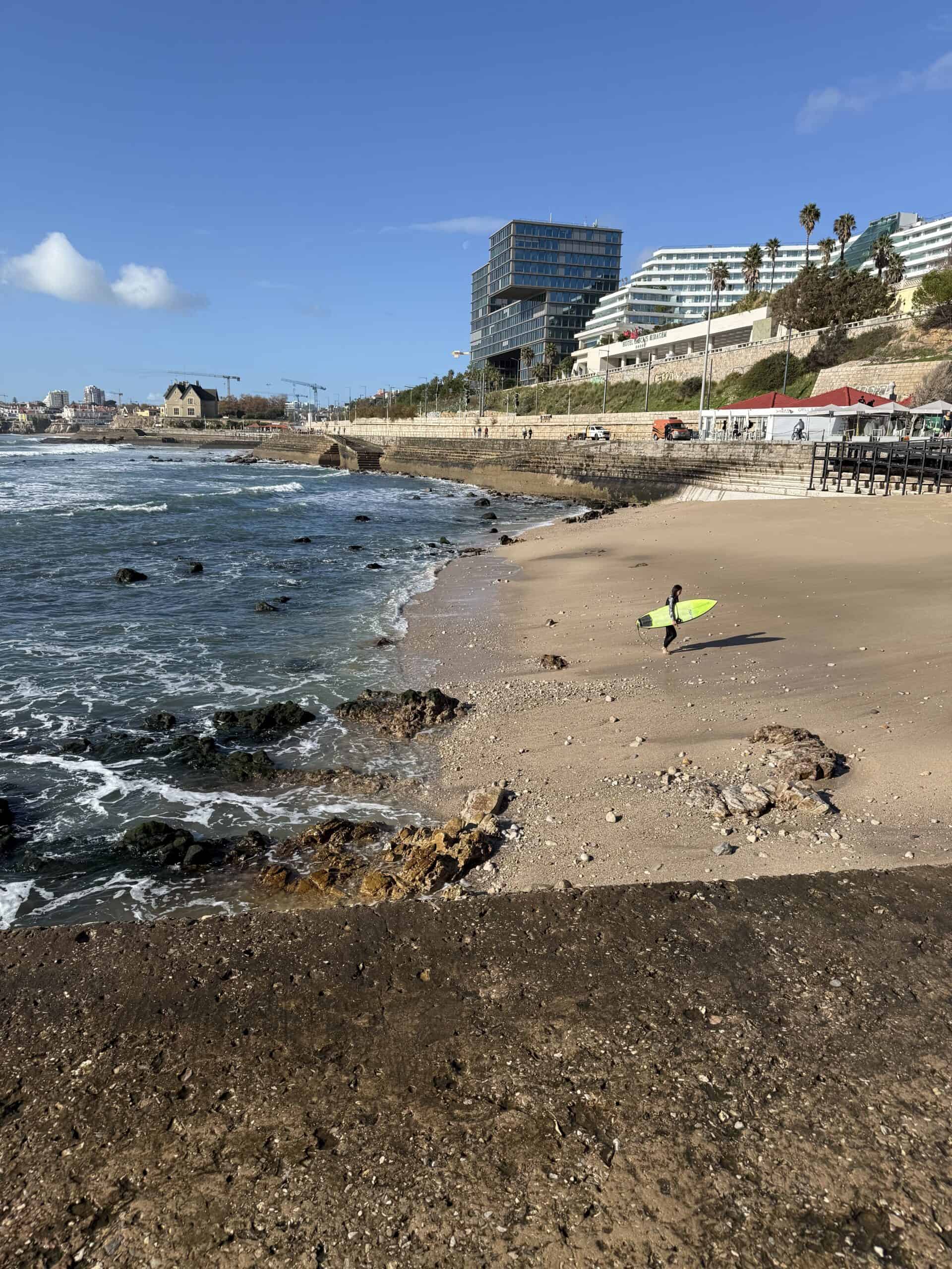 Surfer carrying a bright green surfboard across a sandy beach in Cascais, Portugal, with Atlantic waves and waterfront buildings in the background.