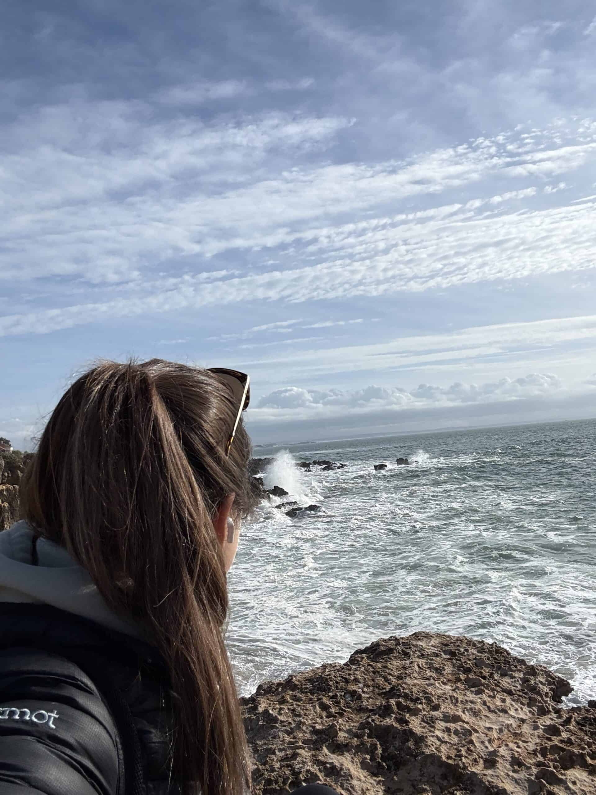 Woman watching Atlantic waves crash against the rocky coastline of Cascais, Portugal, beneath a partly cloudy sky.